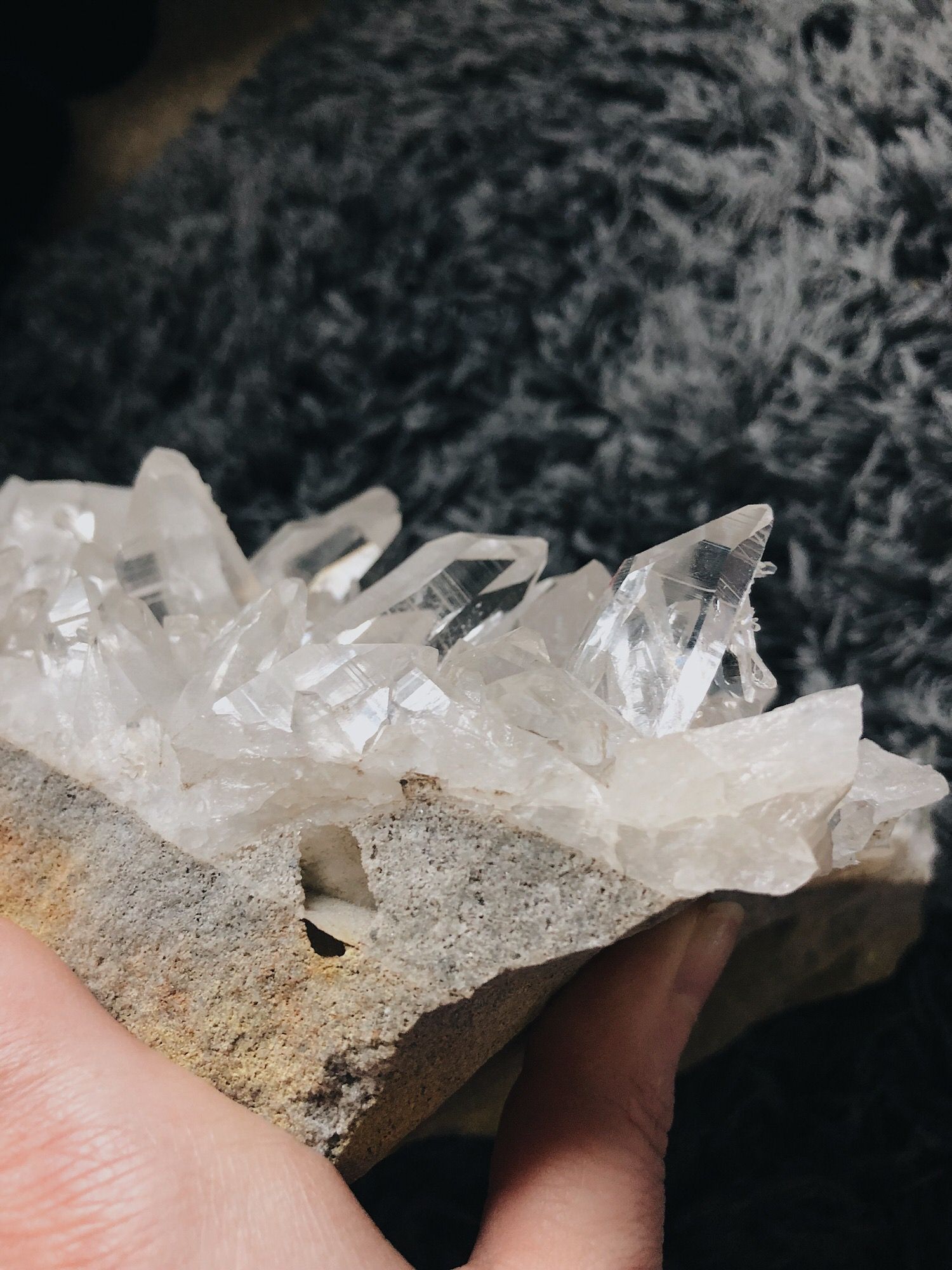 Clear quartz crystal formation growing on a rough stone base, held between fingers showing natural mineral texture.