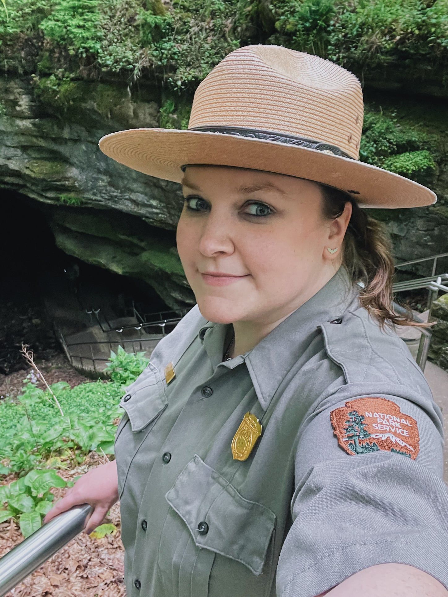 Park ranger wearing a protective sun hat stands by a rocky outcrop in a lush green forest setting.