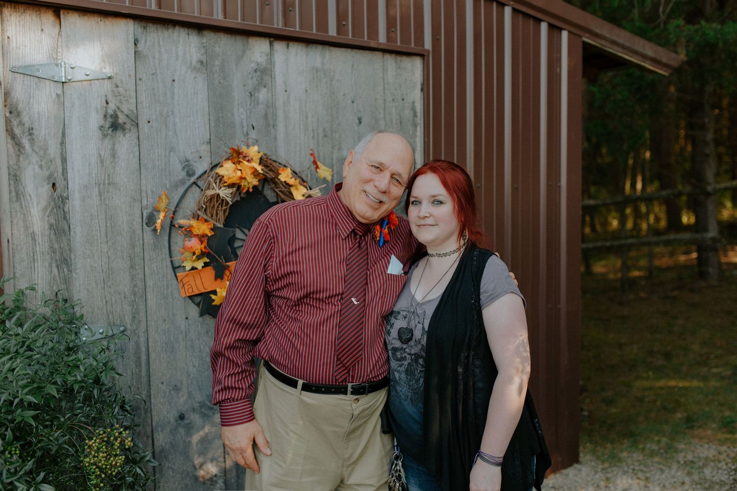 A bald man in a red shirt and a woman with red hair pose together outdoors near a rustic barn with fall wreath.