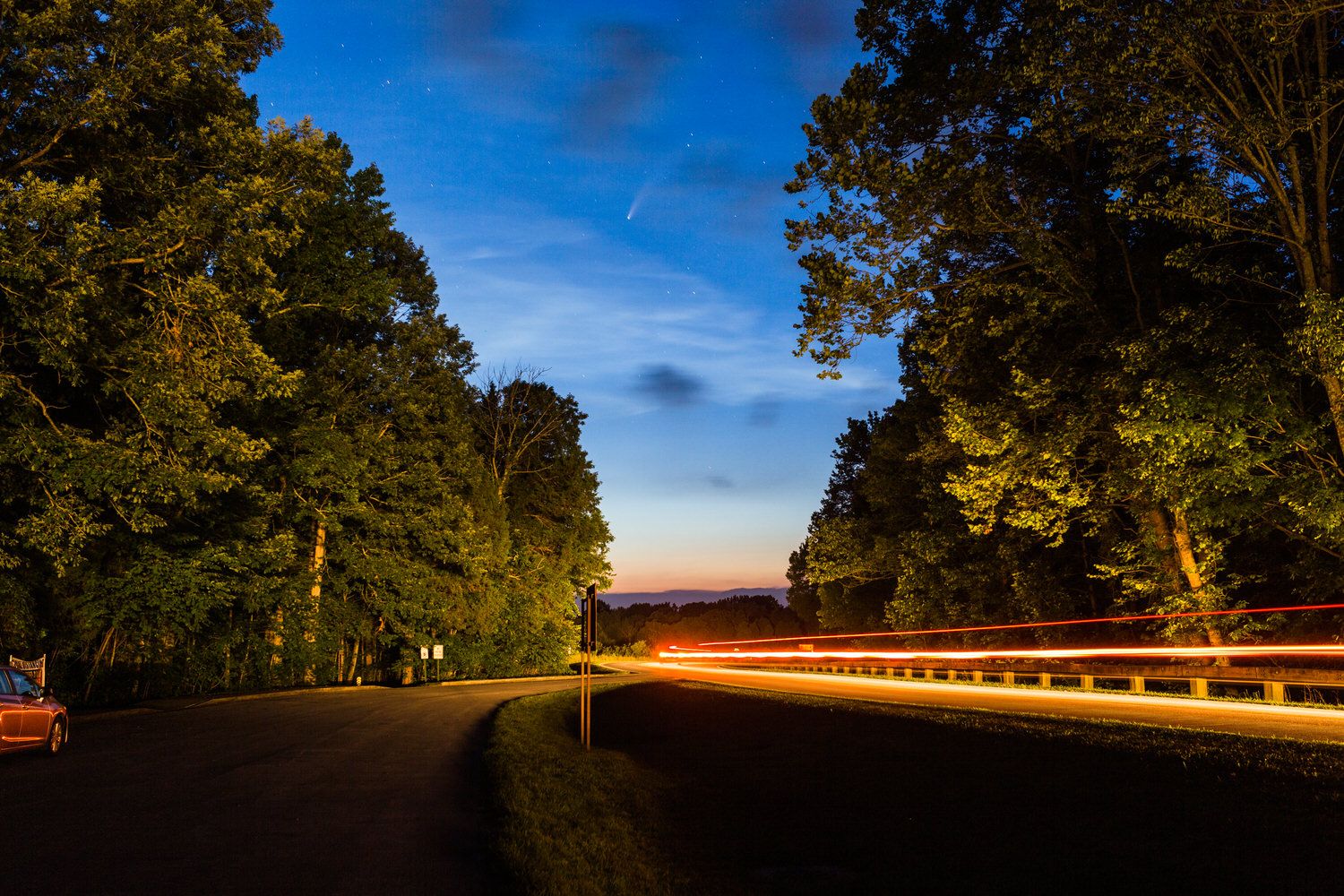 Night road curving through illuminated trees with colorful light trails from passing cars under a twilight blue sky.