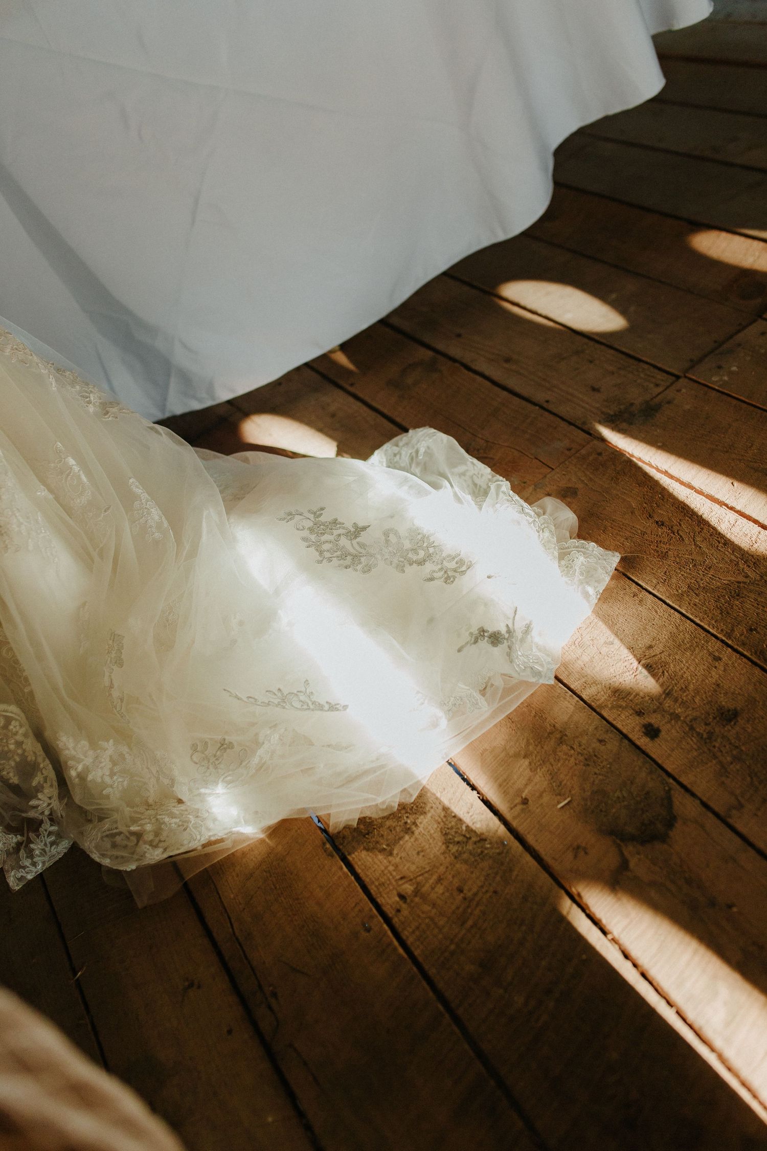 Delicate lace wedding dress train spread on rustic wooden floor bathed in warm golden sunlight.