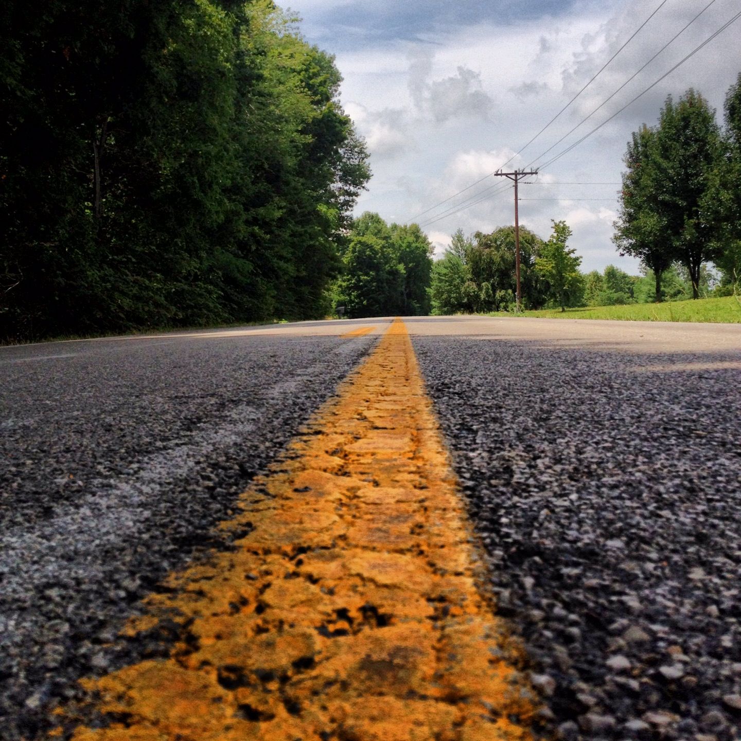 Low-angle view of a two-lane asphalt road with yellow center lines, bordered by lush green trees under a cloudy sky.