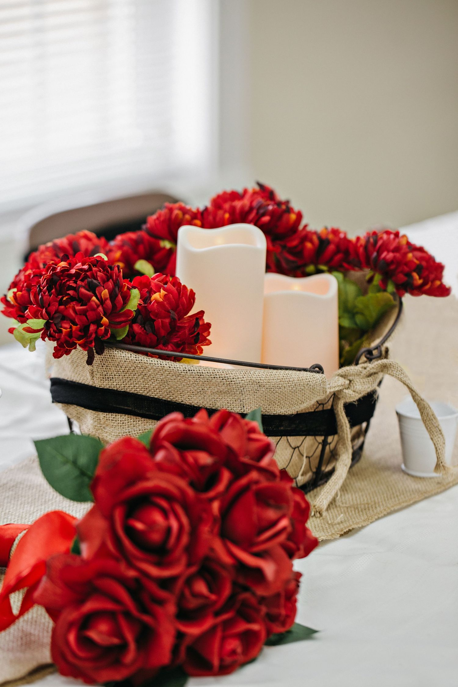 Red chrysanthemums and roses arranged with white pillar candles in a burlap basket centerpiece on a white table.