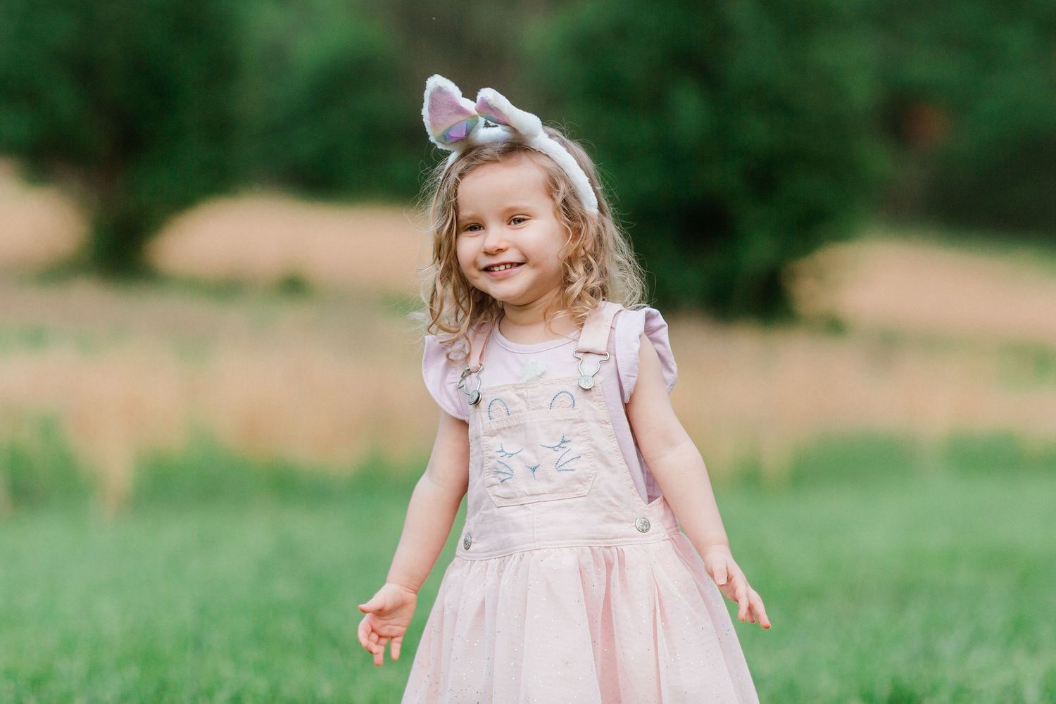 Child in pink tulle dress with bow accessory twirls in grassy outdoor field during summer.