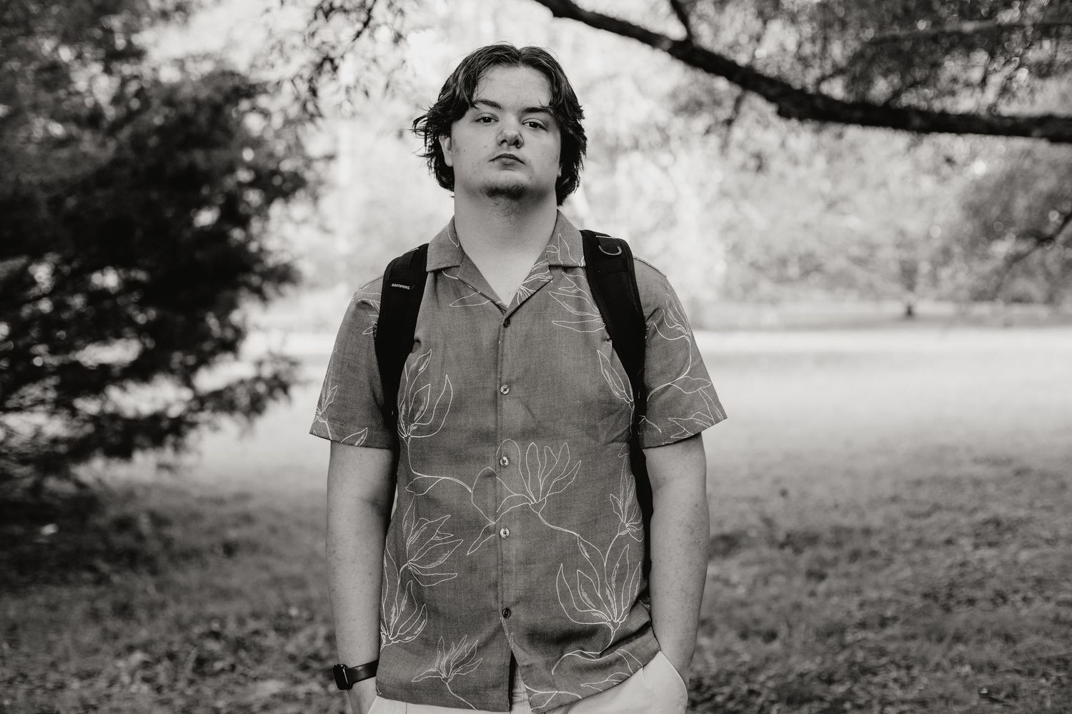 Young man with backpack standing outdoors in park, wearing floral shirt, black and white portrait photo.