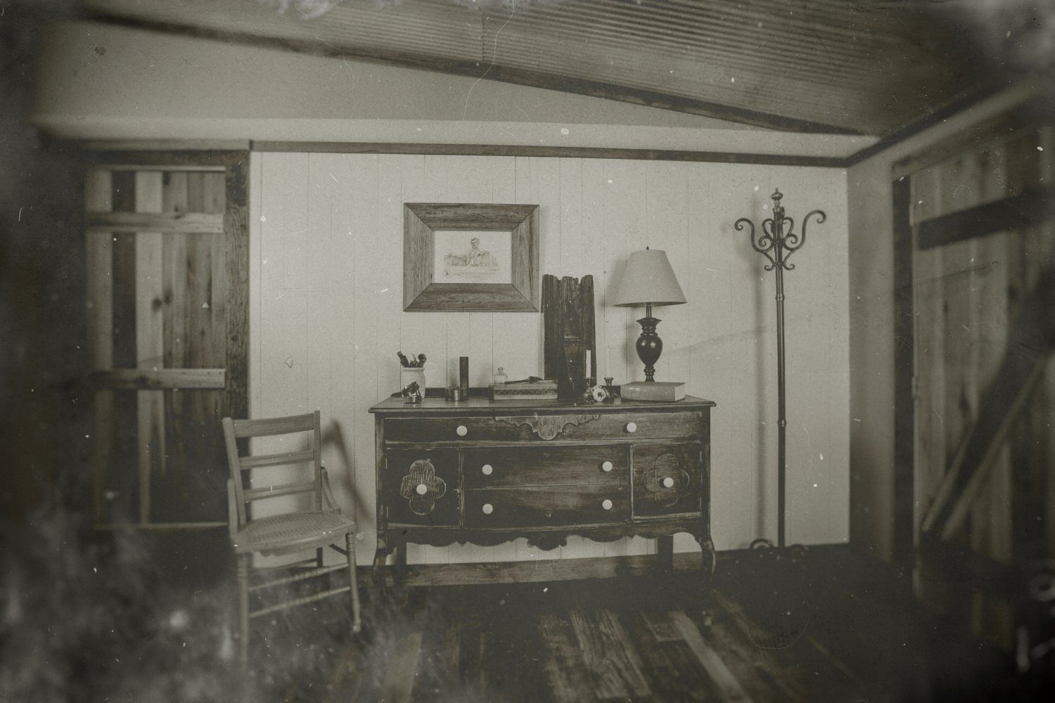 Vintage black and white photo of a rustic room with a wooden dresser, lamp, chair, and framed picture on the wall.
