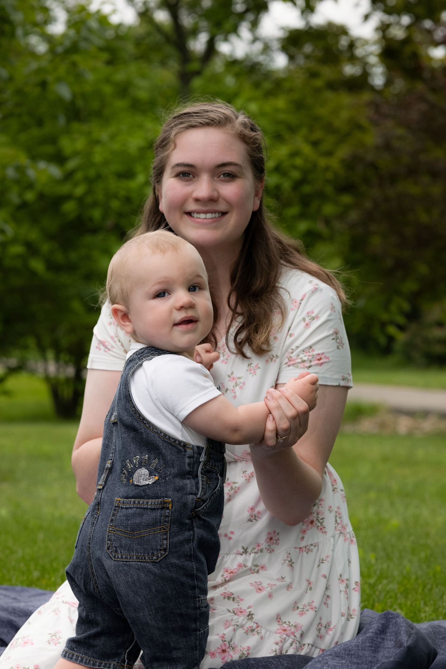 A mother in a white dress lovingly holds her child in a lush green park, captured tenderly by McClure’s Photography.