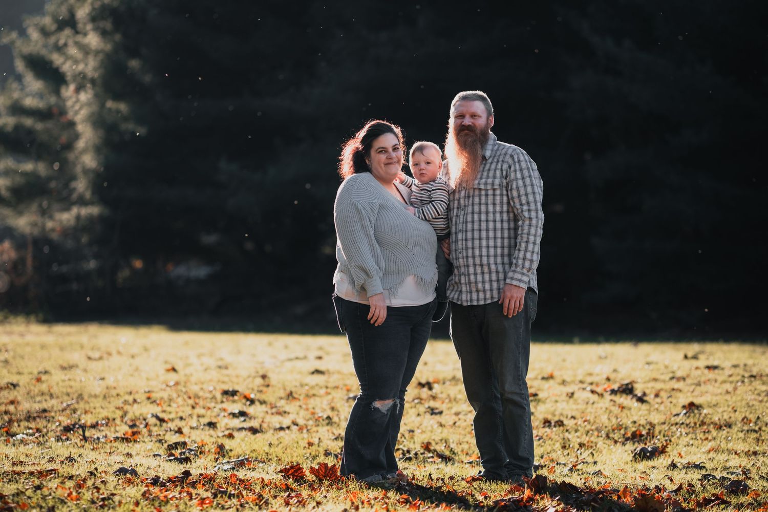A family of three poses in a field, with a woman, man, and child, captured warmly by McClure’s Photography against a dark, wooded backdrop.
