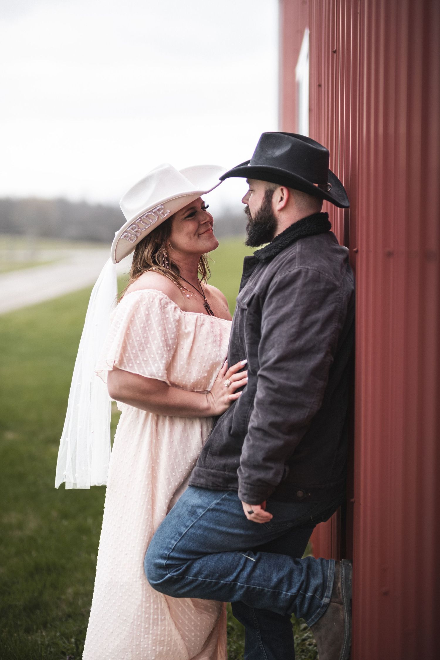 An engaged couple shares a tender moment by a red post, he in a hat and coat, she in a white dress, captured beautifully by McClure’s Photog