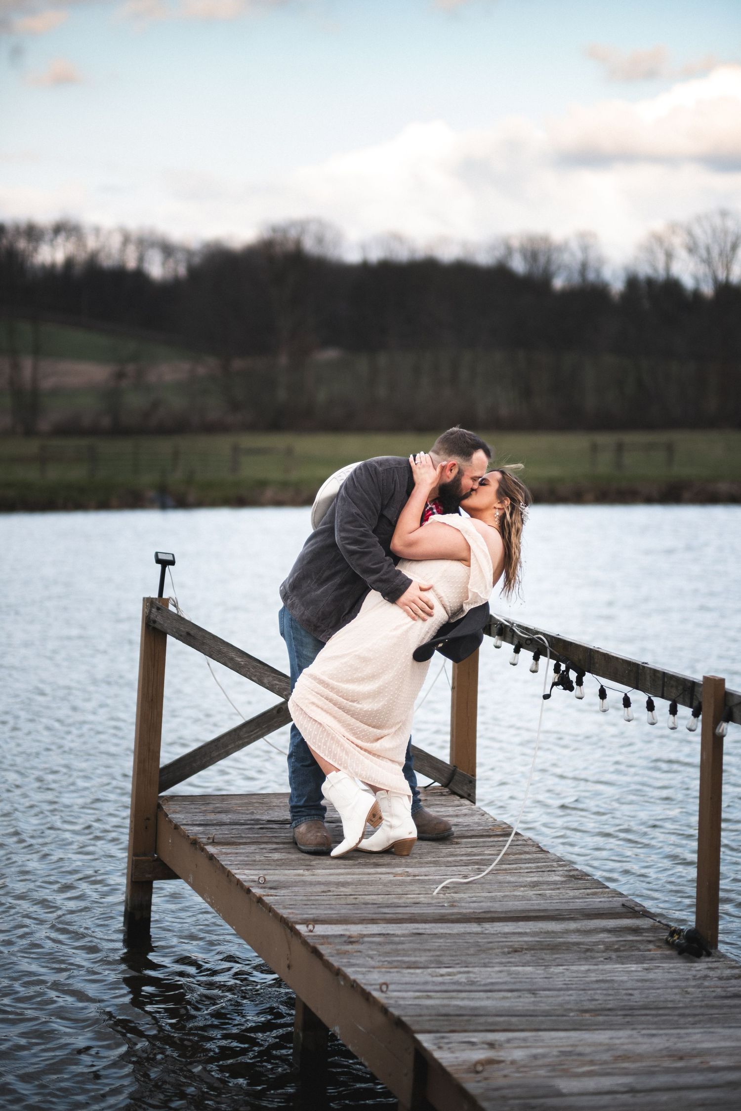 An engaged couple shares a kiss on a wooden dock by the water, surrounded by a serene landscape, captured beautifully by McClure’s Photograp