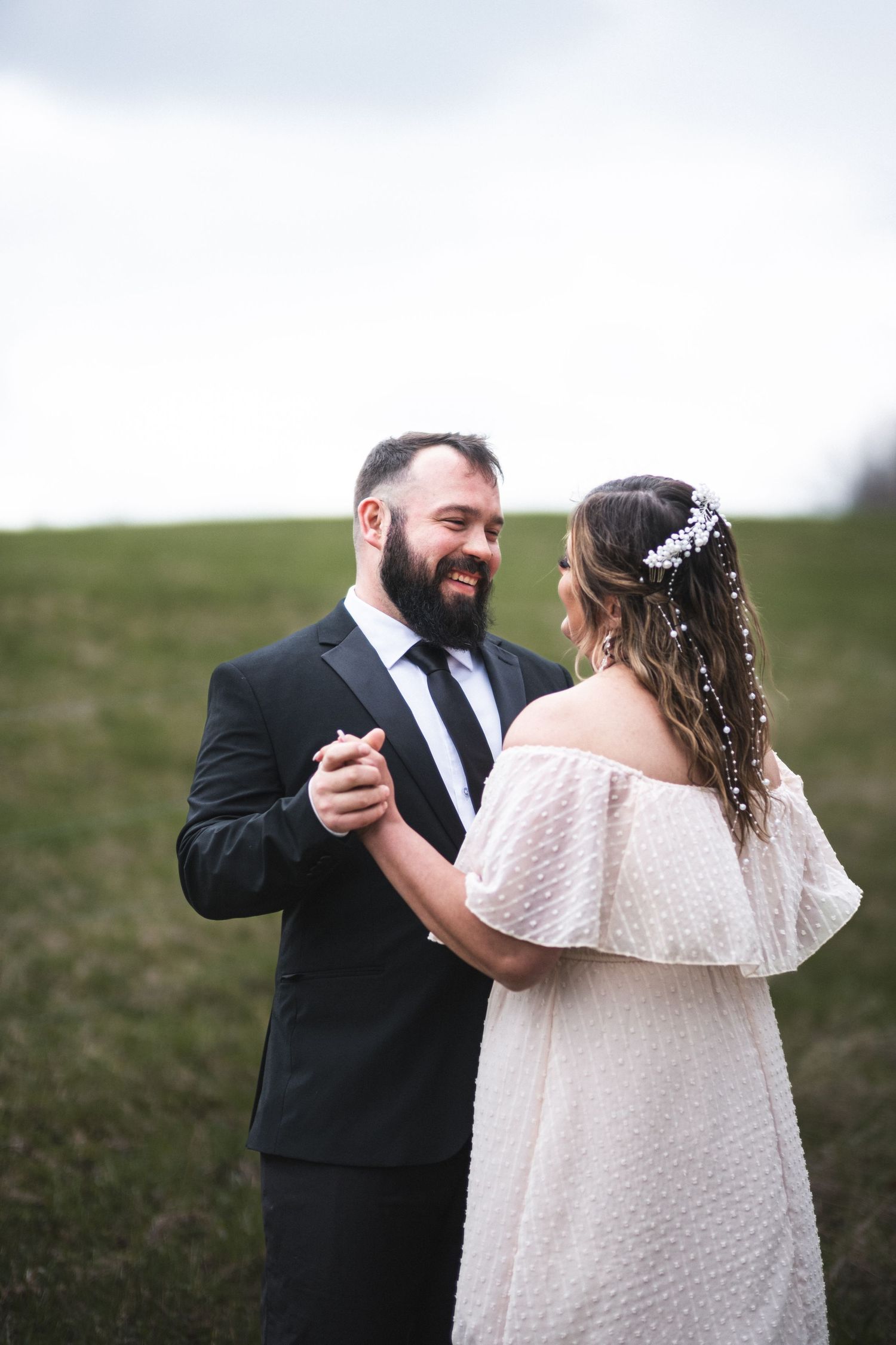 An engaged couple dances in a field, he in a suit, she in a white dress, sharing a joyful moment captured by McClure’s Photography.