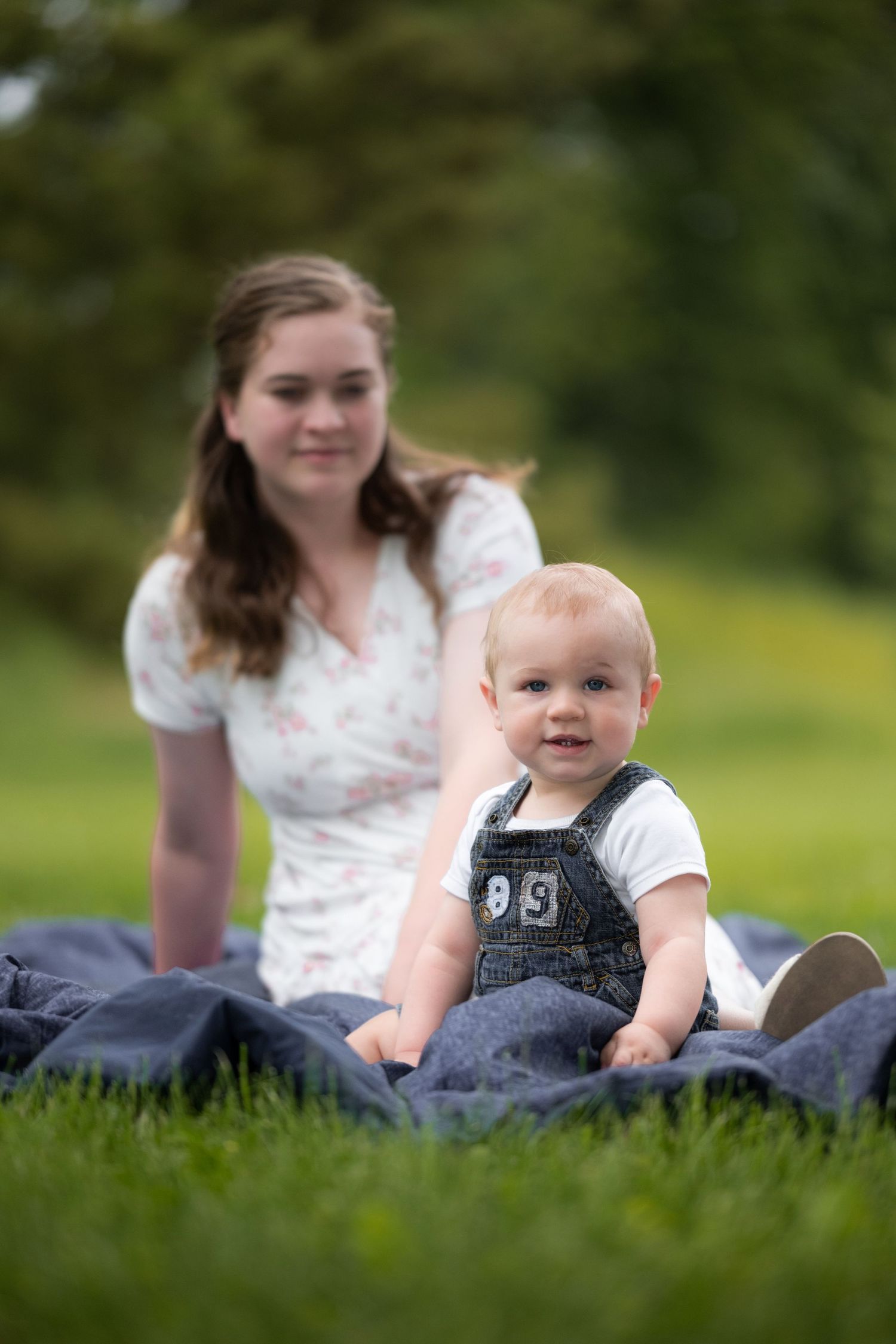 A mother in a white dress lovingly holds her child in a lush green park, captured tenderly by McClure’s Photography.