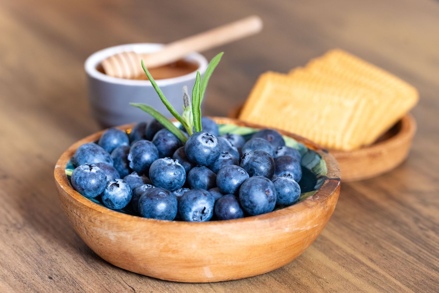 Fresh blueberries in a wooden bowl with crackers and honey on a rustic table.
