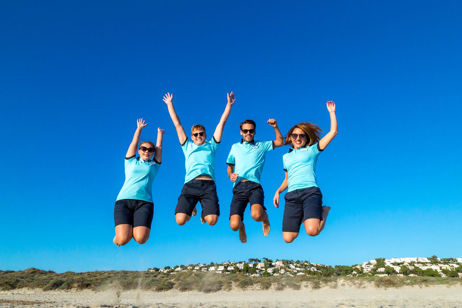 Four people in matching blue shirts jump joyfully together on a sandy beach against a bright blue sky.