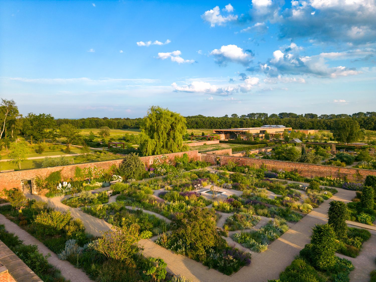 Aerial view of a formal garden with organized flower beds and paths under a bright blue summer sky with fluffy clouds.