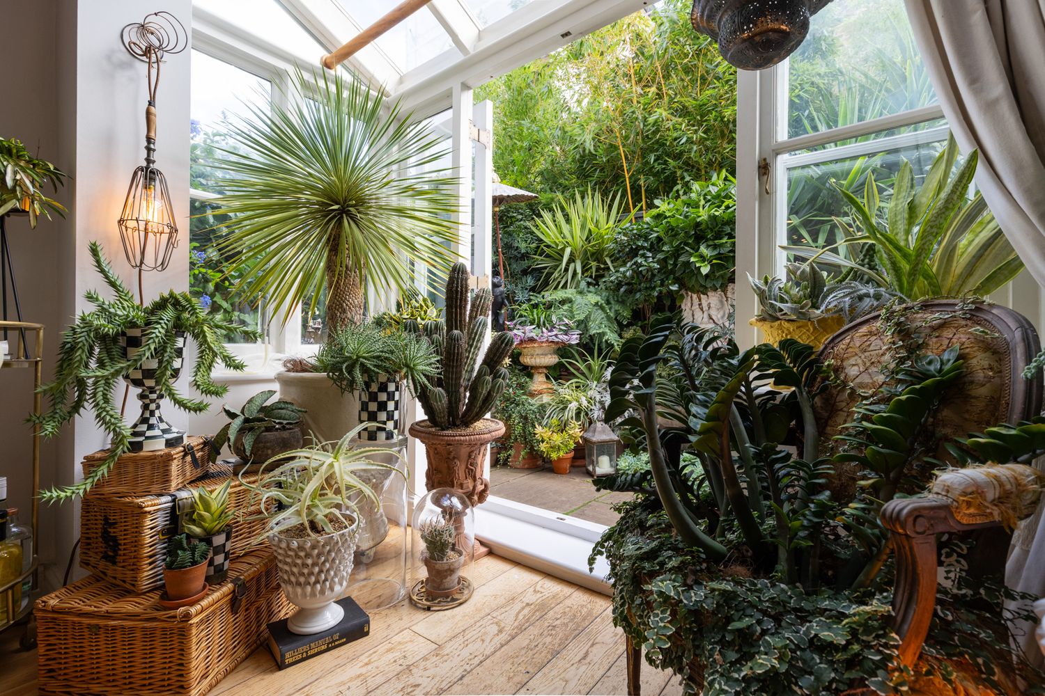 A bright sunroom filled with potted tropical plants and decorative ornaments sits beneath a skylit conservatory ceiling.