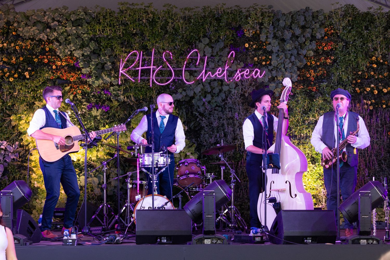 A live band performs on stage at night with instruments in front of a floral wall and neon signage.