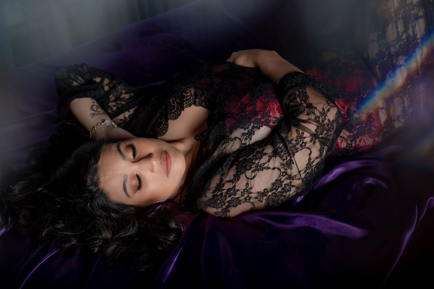 A figure wearing black lace reclines on purple fabric in a dark and moody boudoir setting.