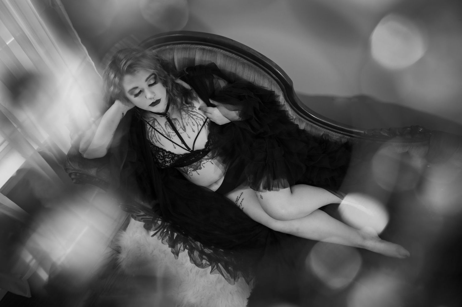 Dreamy black and white boudoir photo of a figure reclining on dark furniture with soft bokeh effects.