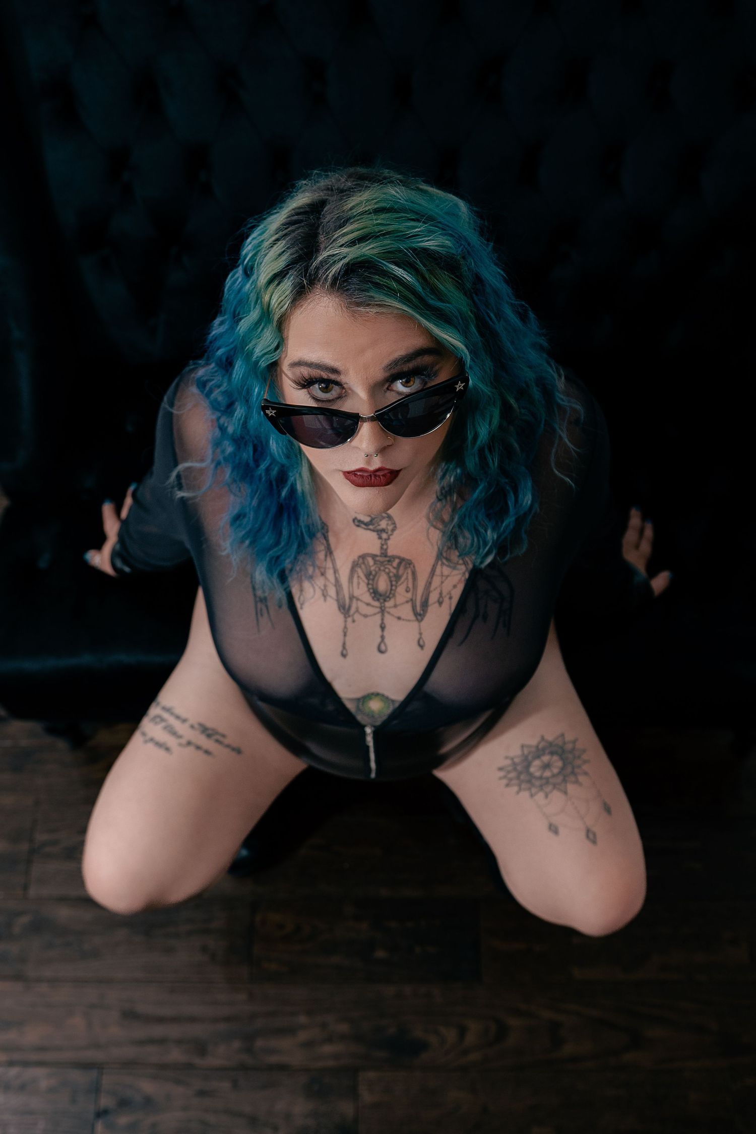 Alternative model in sunglasses and dark mesh top poses on wooden floor with vibrant blue-green hair.