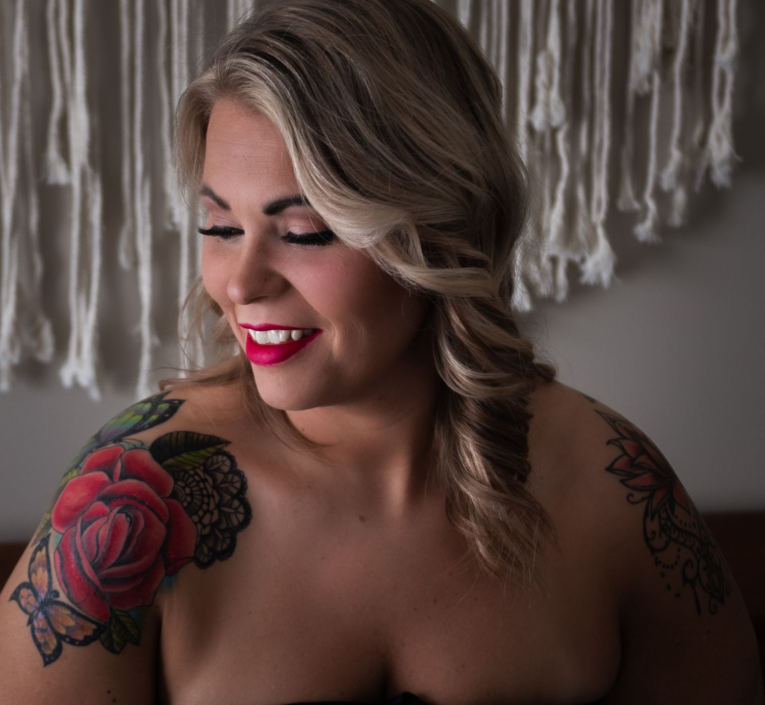 A woman with blonde wavy hair, red lipstick and floral arm tattoos smiles with closed eyes in front of macrame decor.