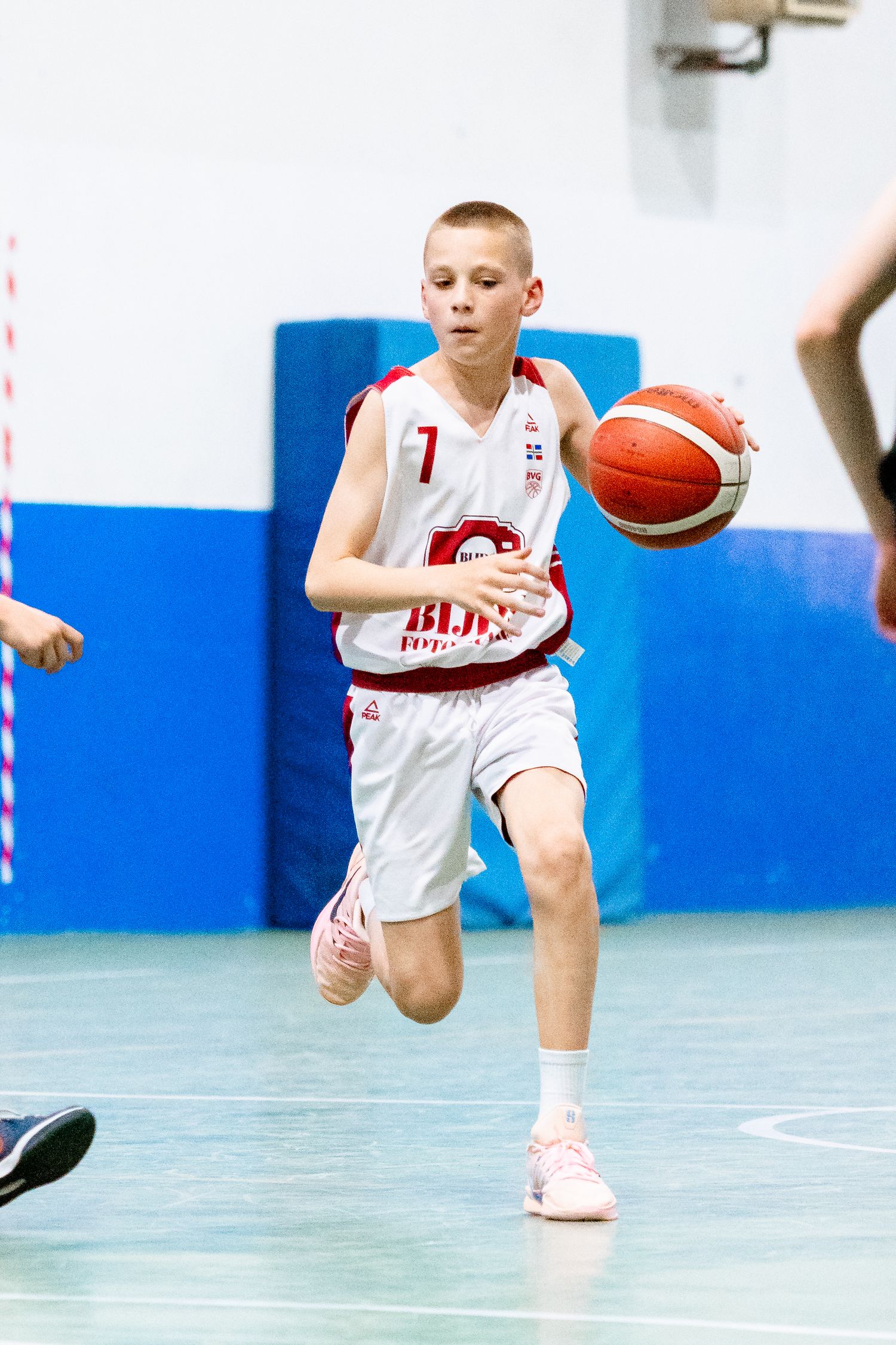 Young boy in white basketball uniform dribbling ball during youth league game on indoor court.