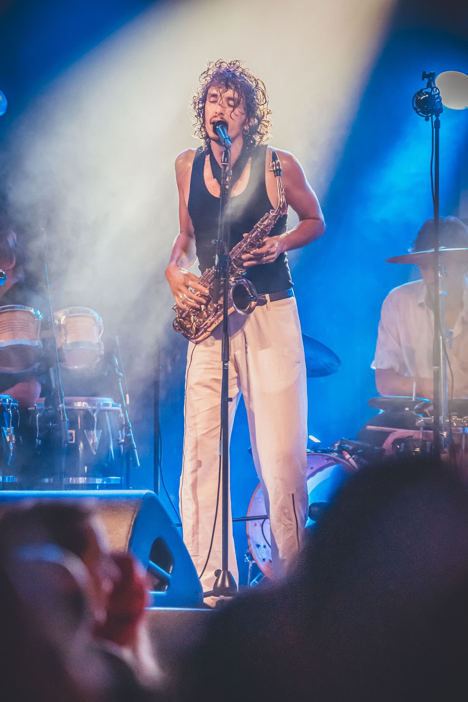 Musician performs with saxophone on stage under dramatic blue and white spotlights with percussion instruments in background.