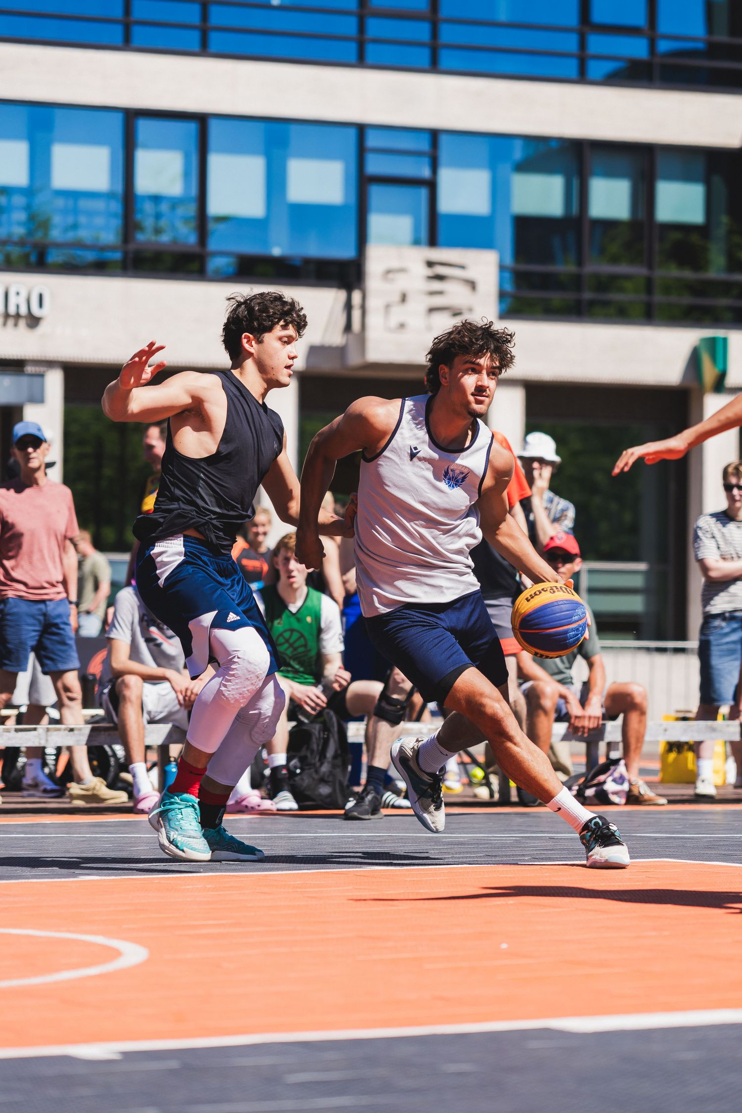 Two young men compete in an outdoor 3x3 basketball game, one dribbling past a defender before a crowd.