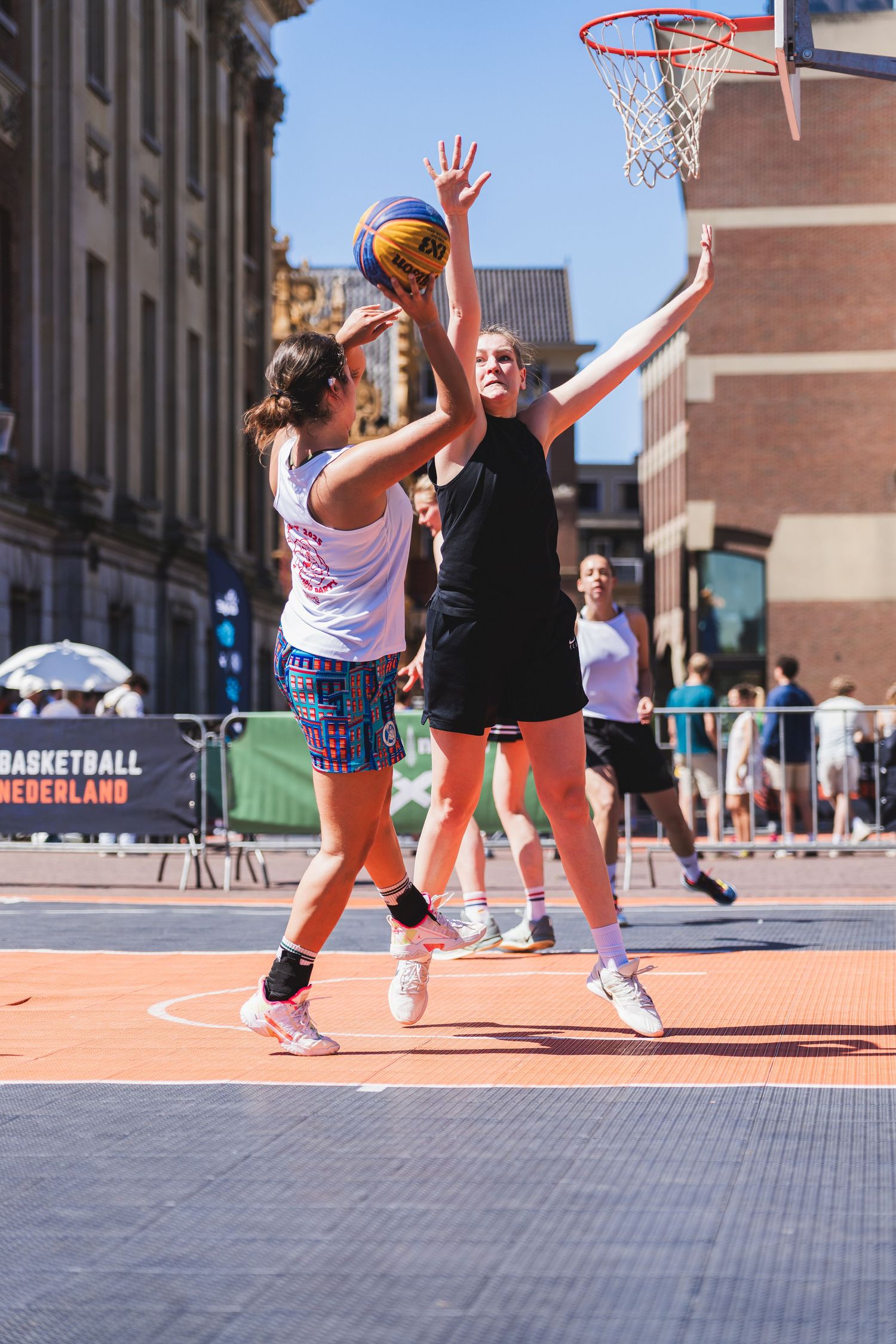 Two women play 3x3 street basketball outdoors, one shooting near the hoop as the other defends on a sunny day.