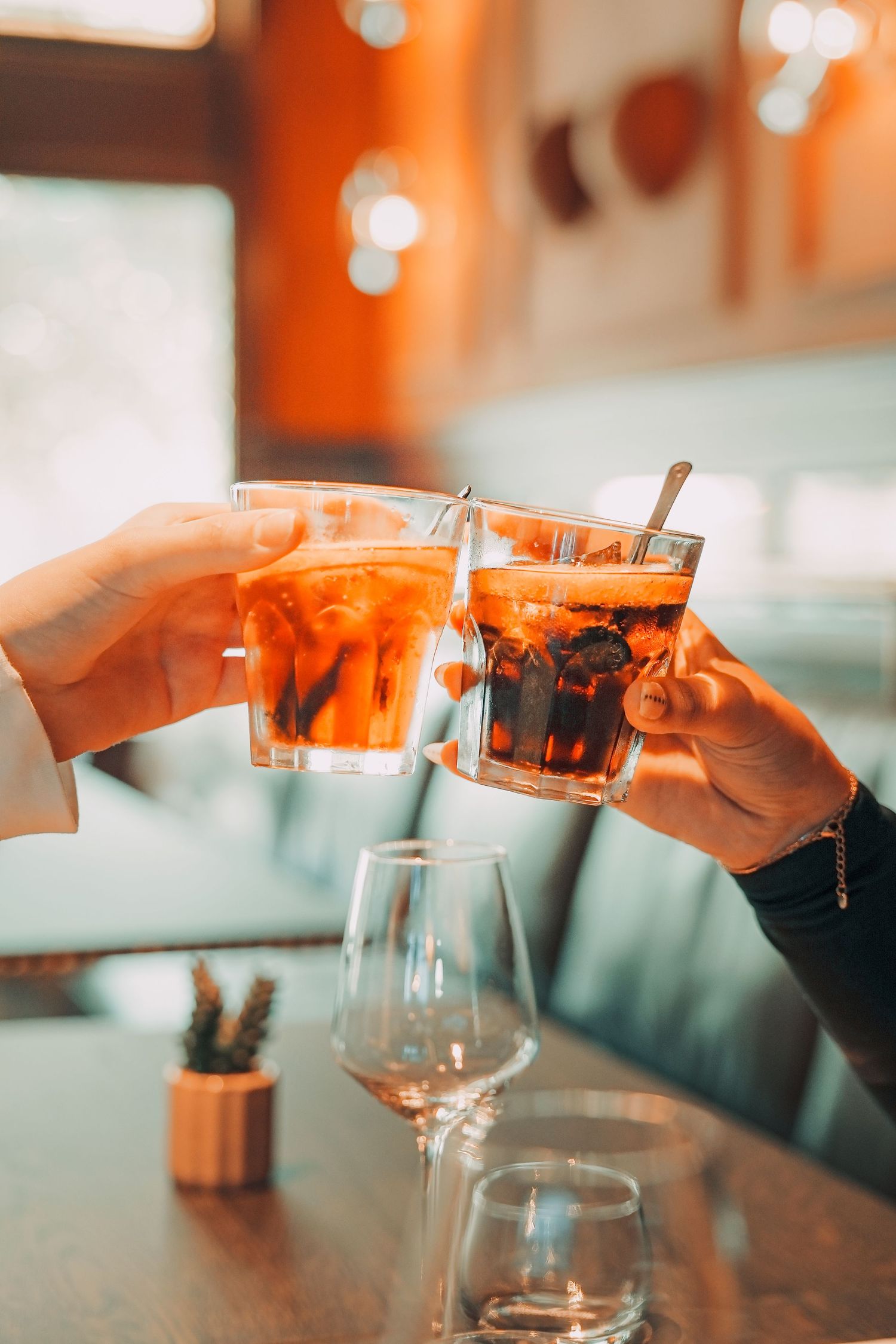 Two hands clinking glasses of iced cocktails at a restaurant table, toasting with amber drinks.