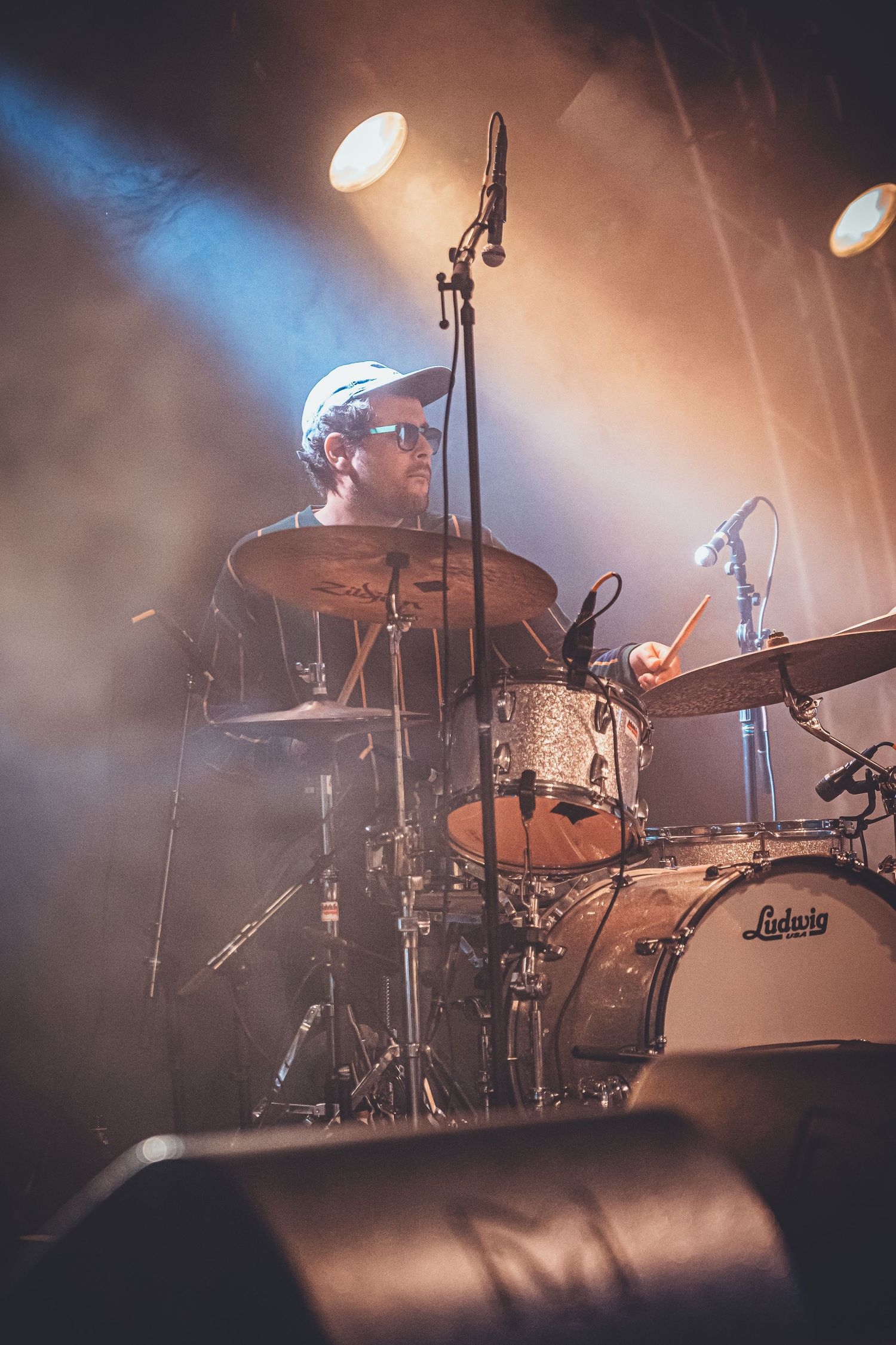 A drummer performs on stage behind a Ludwig drumkit with dramatic blue stage lighting and smoke effects in the background.