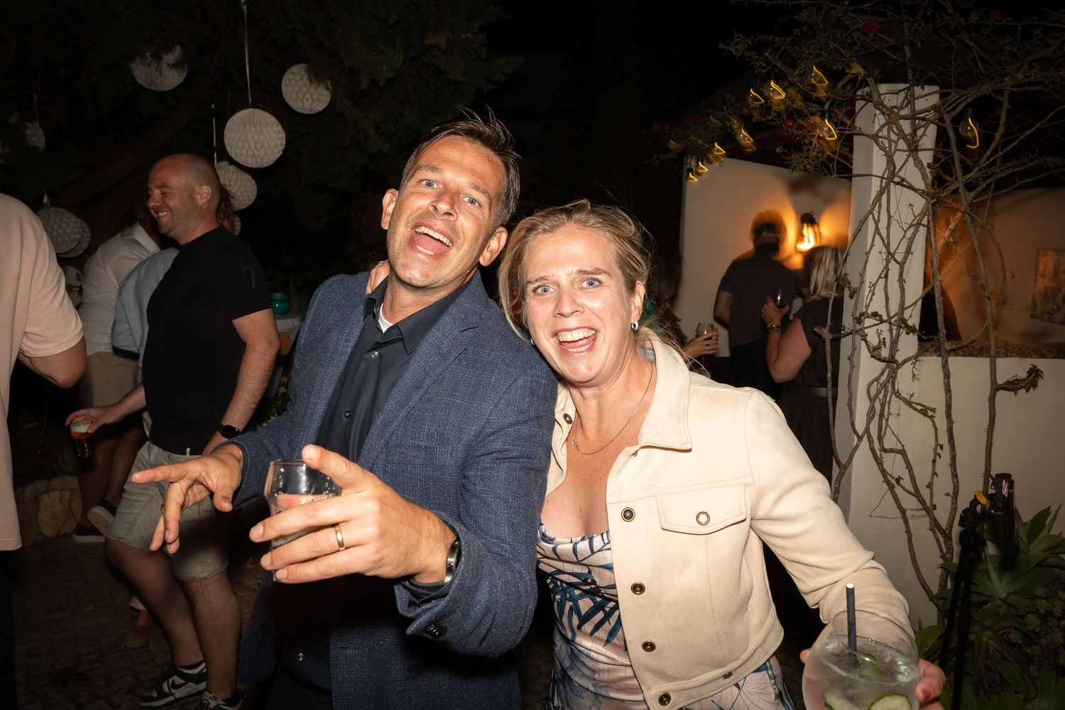 A couple laughs together during a nighttime outdoor party with decorative lights and a festive atmosphere.