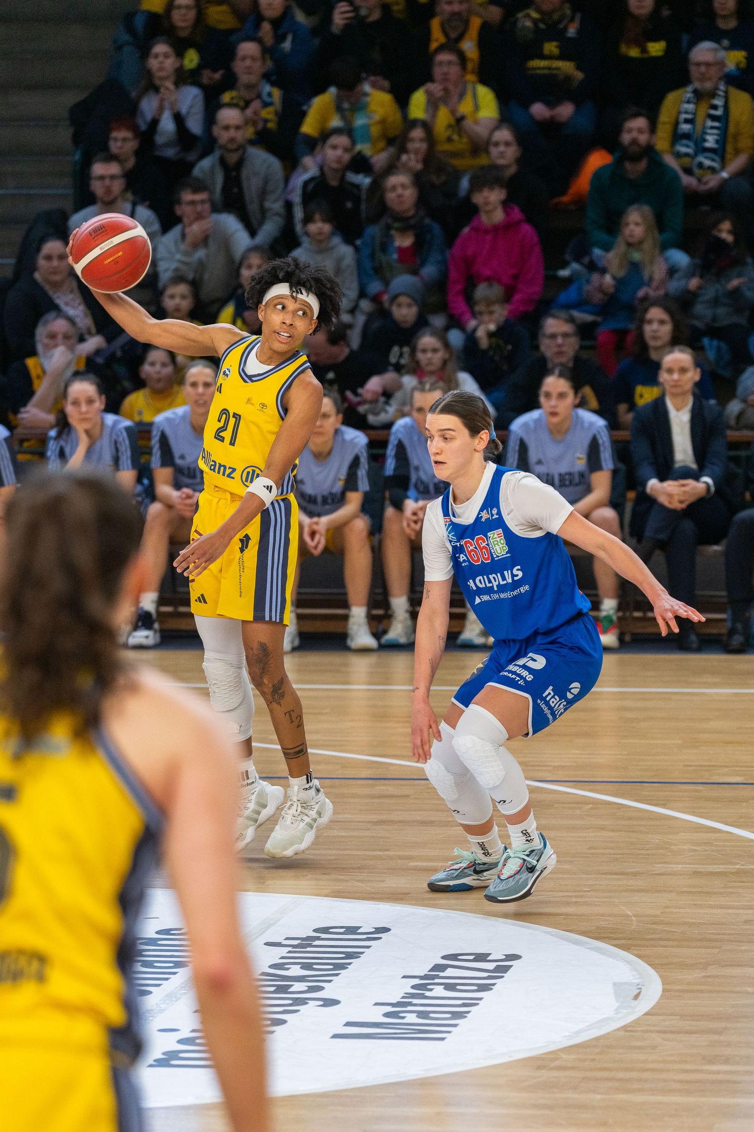 Women's basketball game action with player #21 in yellow reaching for ball while blue team defender guards closely.