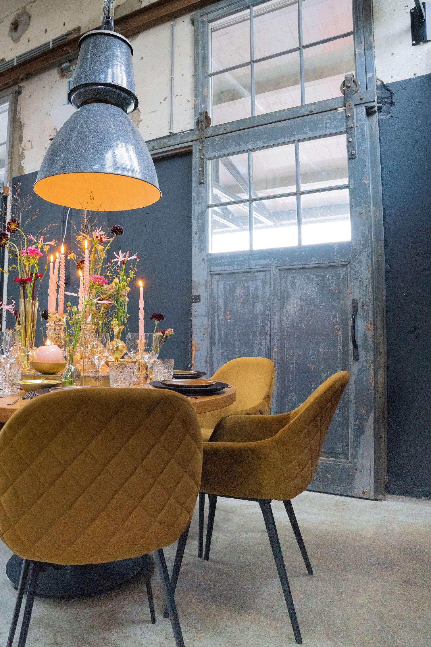Industrial dining space with yellow velvet chairs and pendant lamp over wooden table with flowers.
