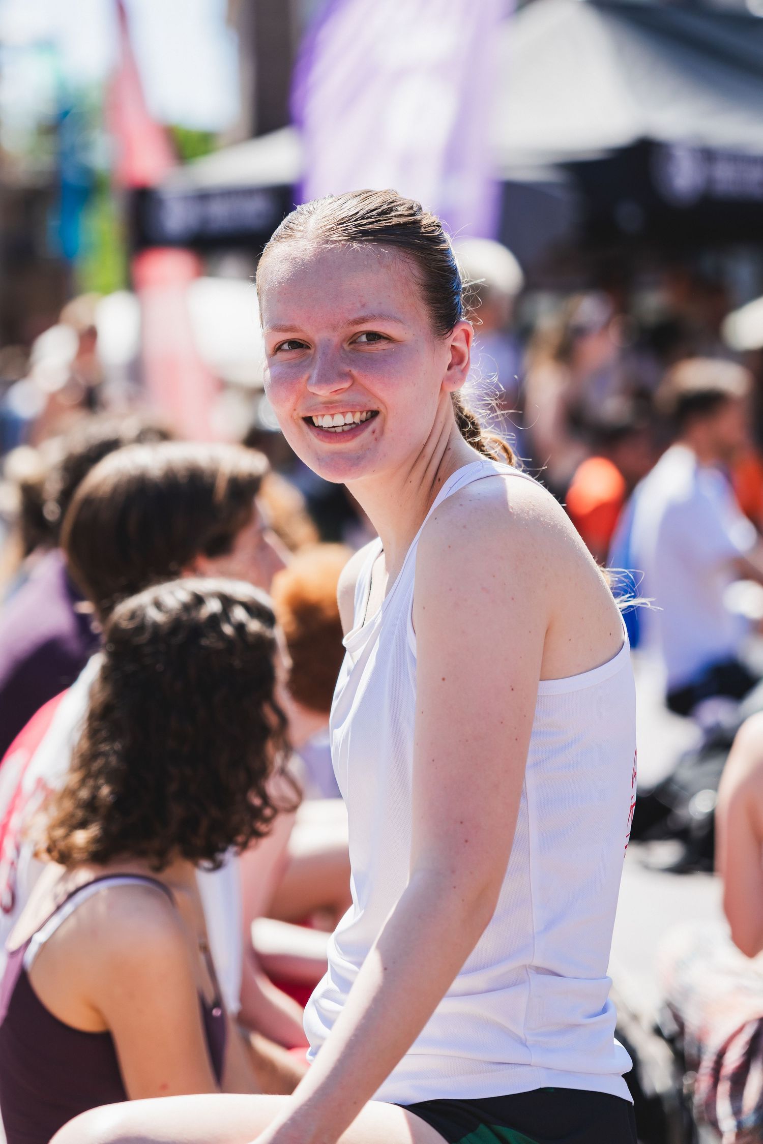 Smiling young woman in white tank top sits among crowd at an outdoor summer basketball tournament.