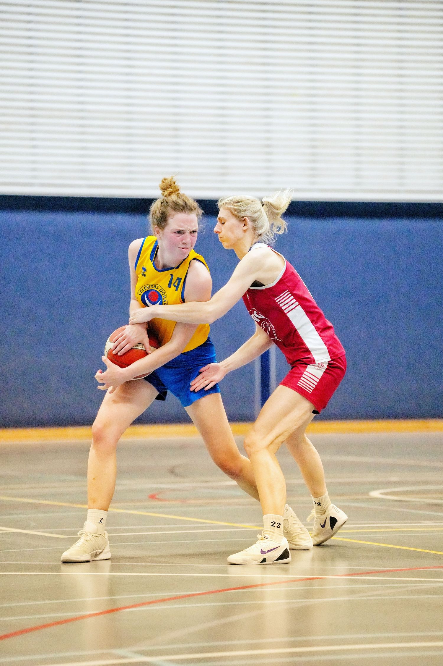 Two female basketball players compete during an indoor game, one in yellow defending the ball against a red-uniformed opponent.