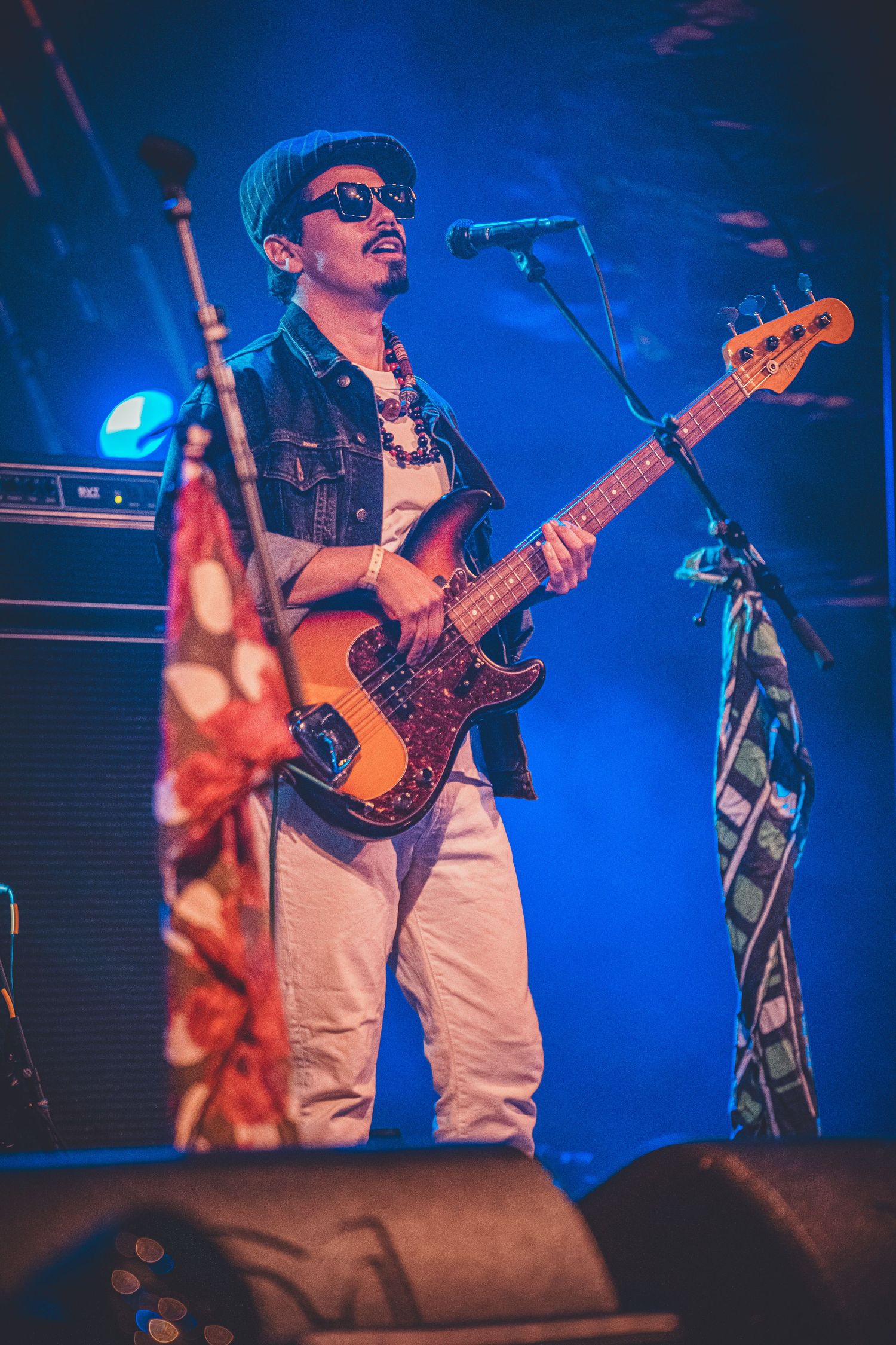 Bass guitarist performs on stage against vibrant blue lighting backdrop wearing sunglasses.