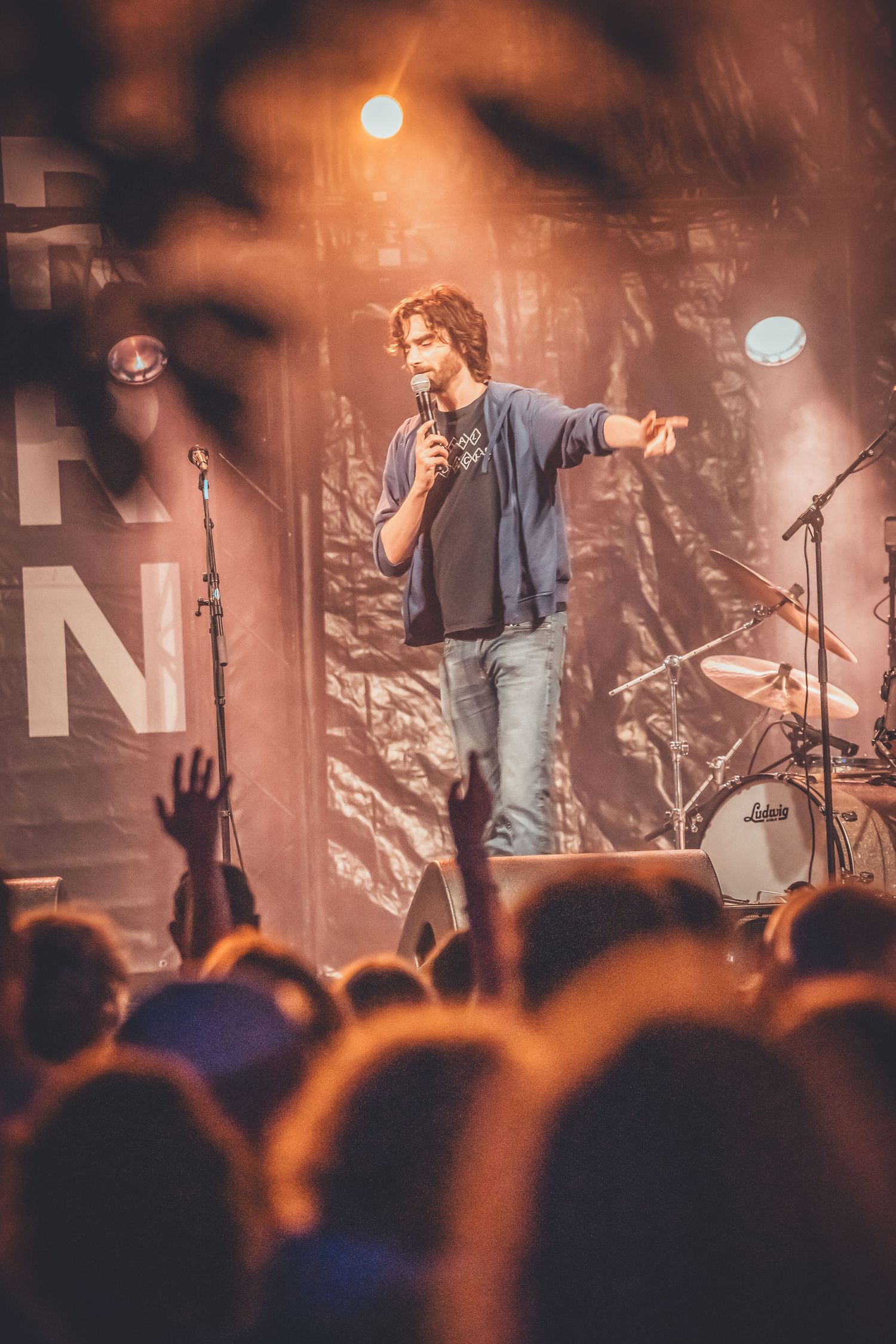A performer on stage with guitar sings passionately to an energetic crowd at a dimly lit concert venue.