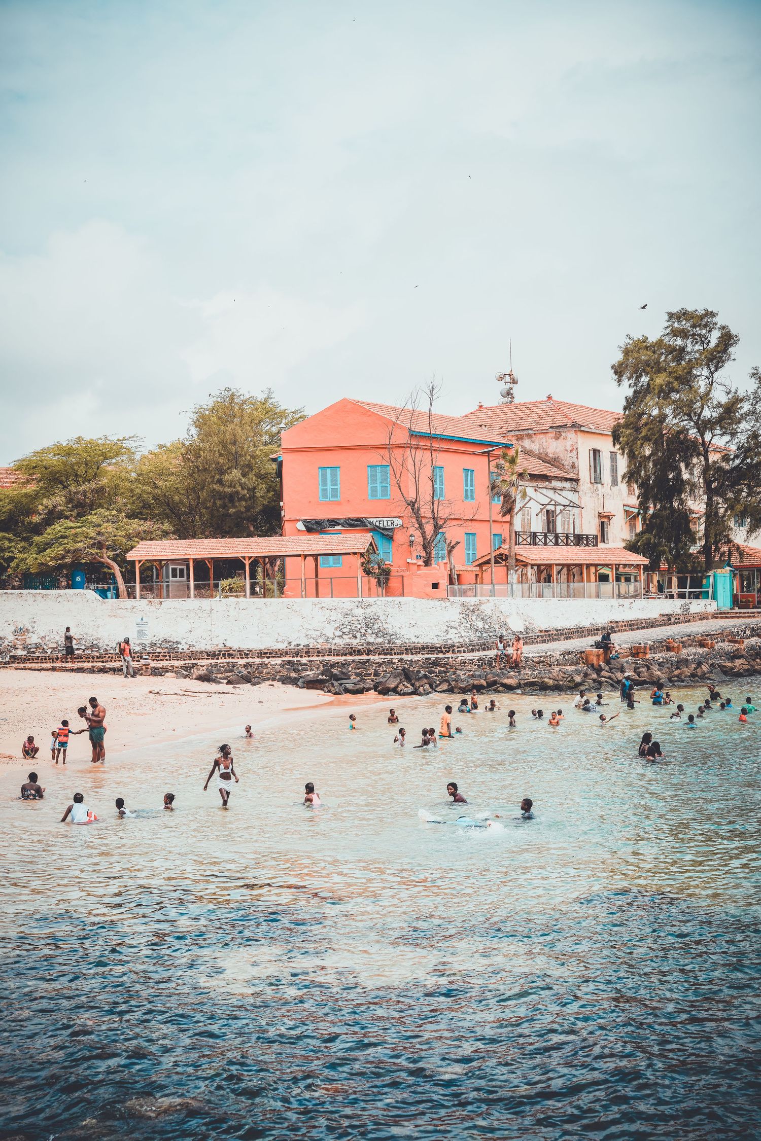 People swimming near a vibrant coral-colored colonial building on the shores of Goree Island, Senegal.