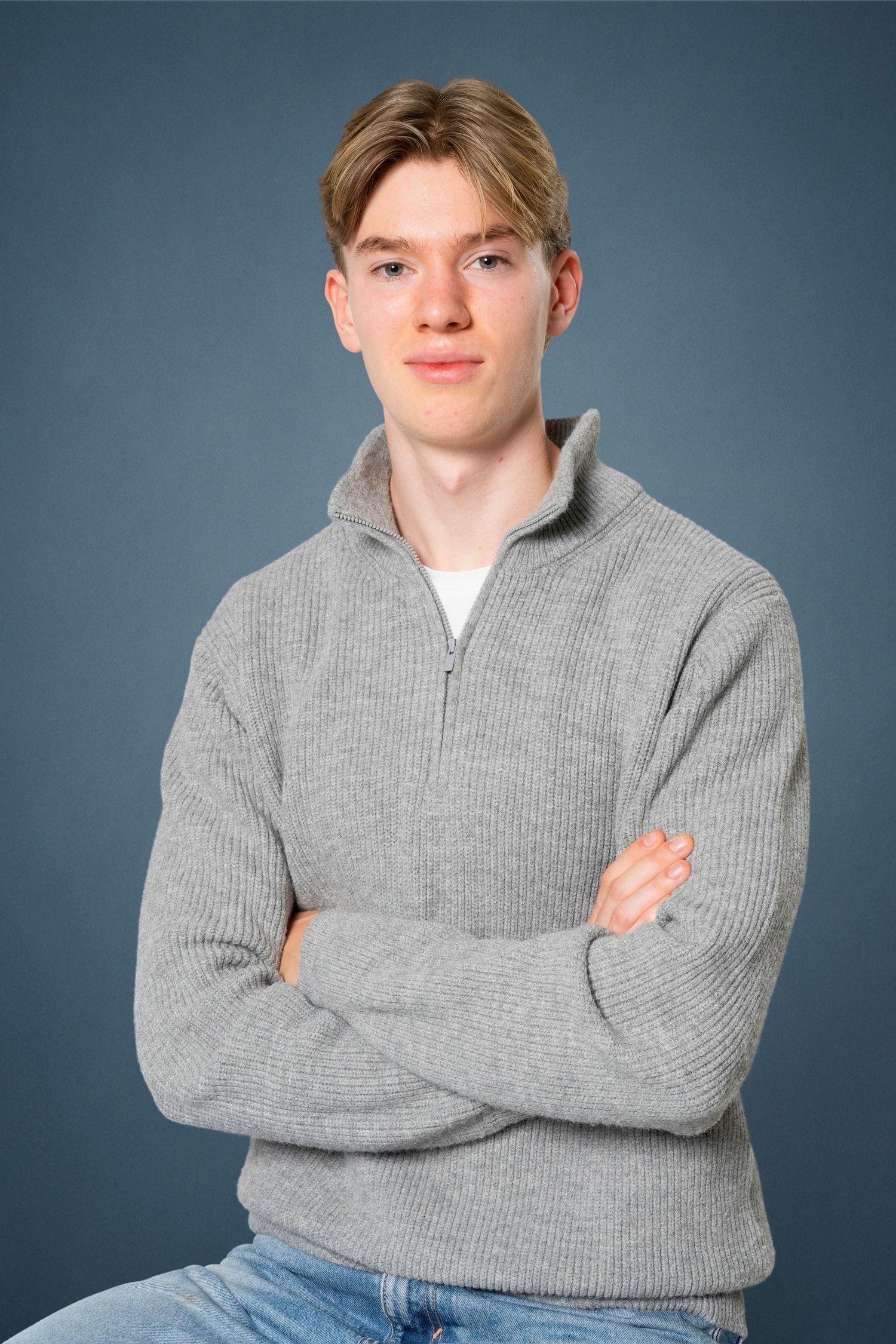 Young man with short blond hair sits with arms crossed, wearing a gray quarter-zip knit sweater and jeans against a blue studio backdrop.