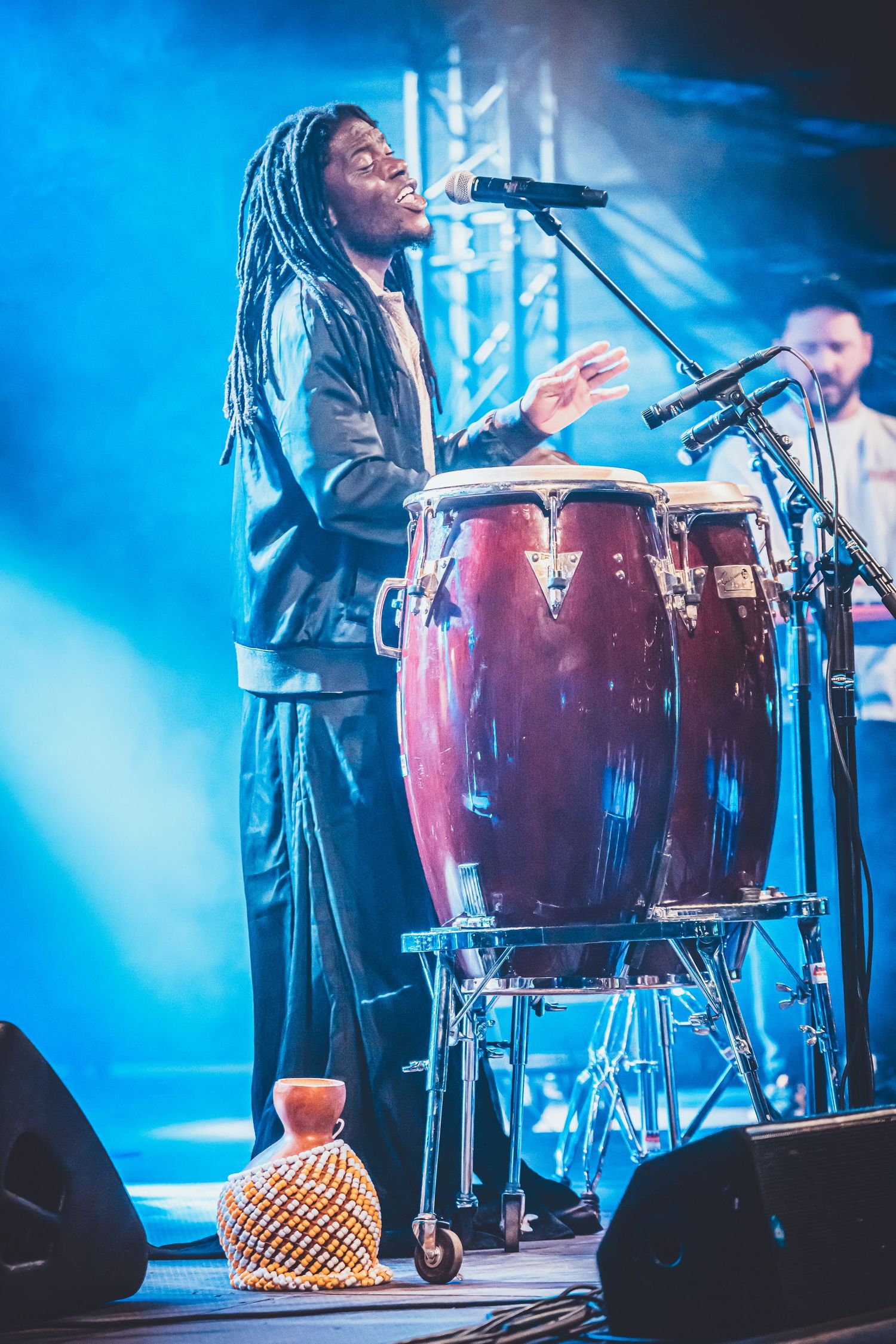 A musician performs with a drum setup on stage surrounded by blue atmospheric lighting during a live concert performance.