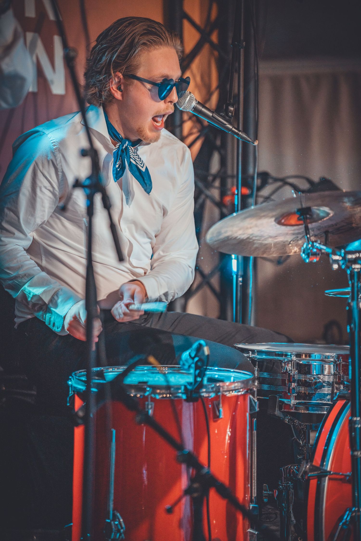 Drummer performs energetically behind red drum kit with blue stage lighting effects.