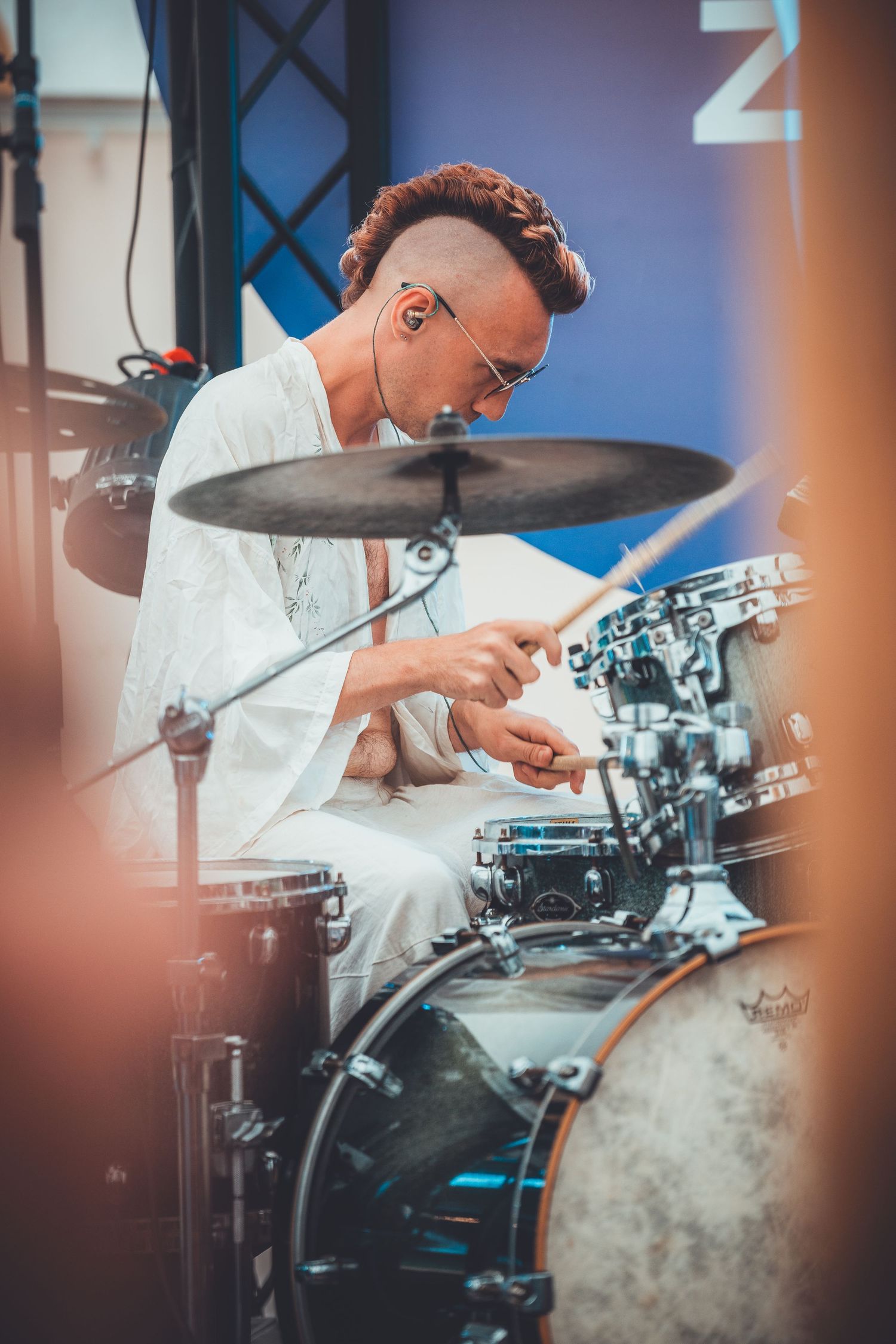 A drummer in a white shirt intensely plays a drum set during a performance with stage lighting effects.