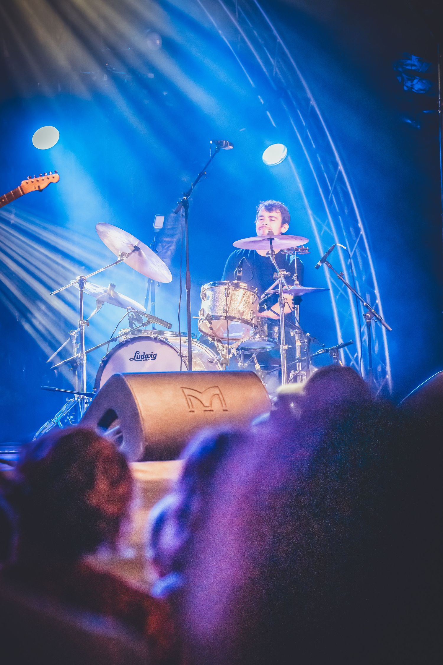 A concert stage illuminated in vibrant blue lighting shows a drum kit setup with beams of light cutting through the atmosphere.