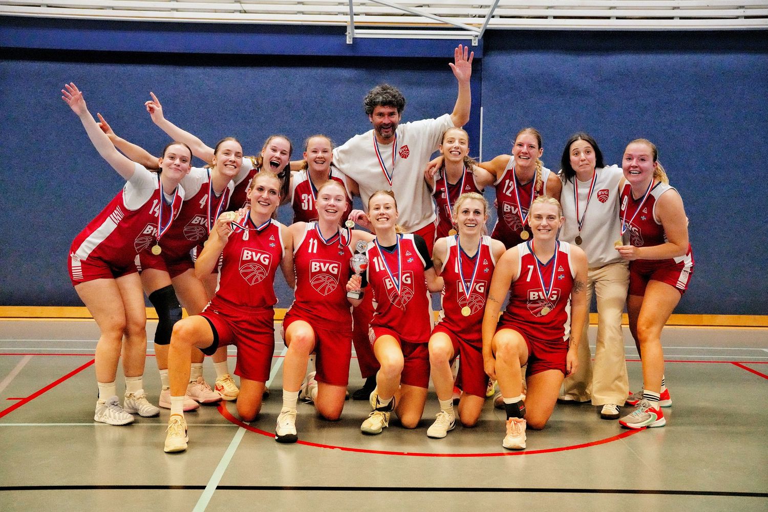 Girls volleyball team in red uniforms celebrating championship win with medals on indoor court.