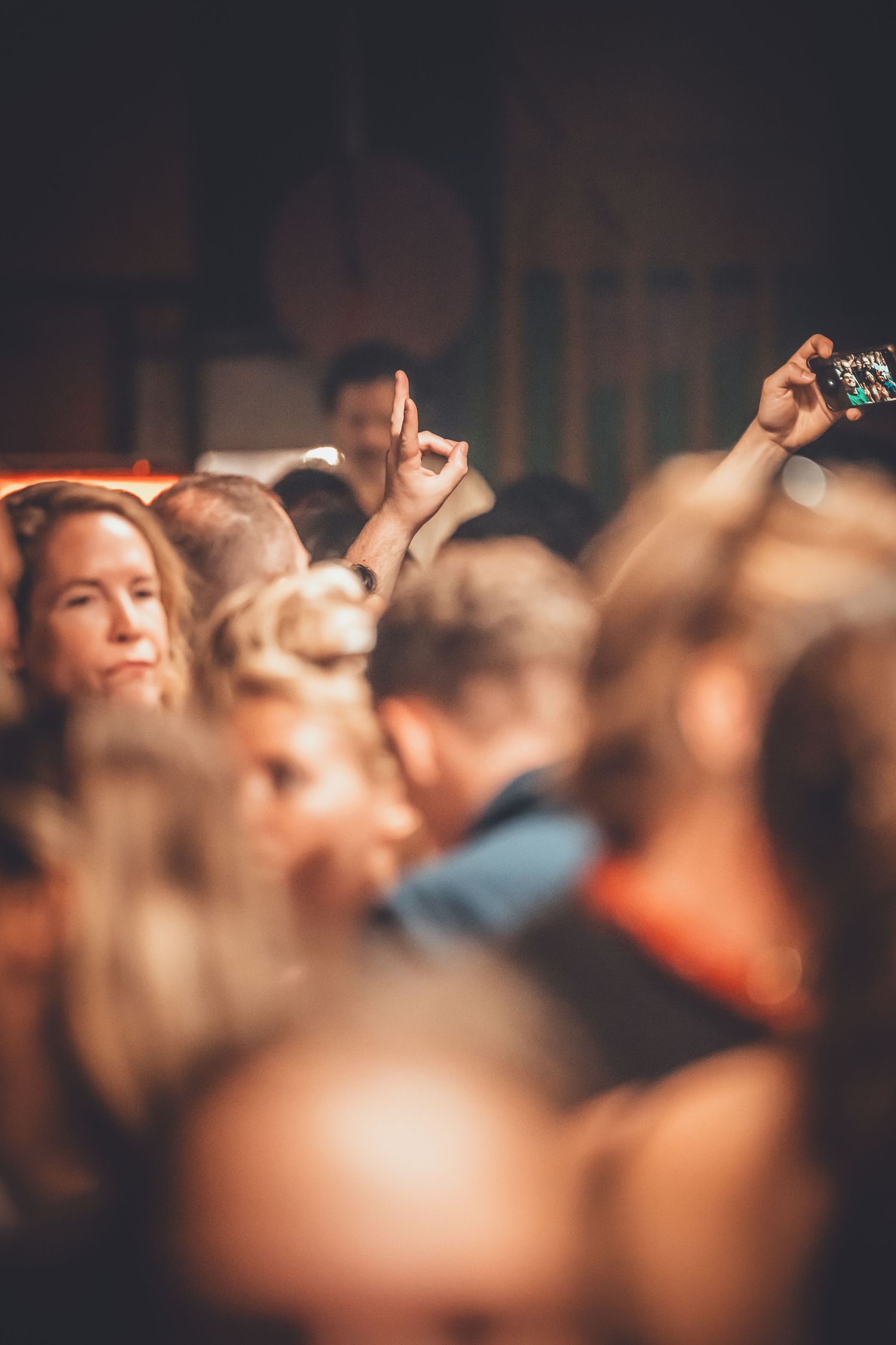 Crowd of people at a concert or event with raised hands and phones in a dimly lit venue.