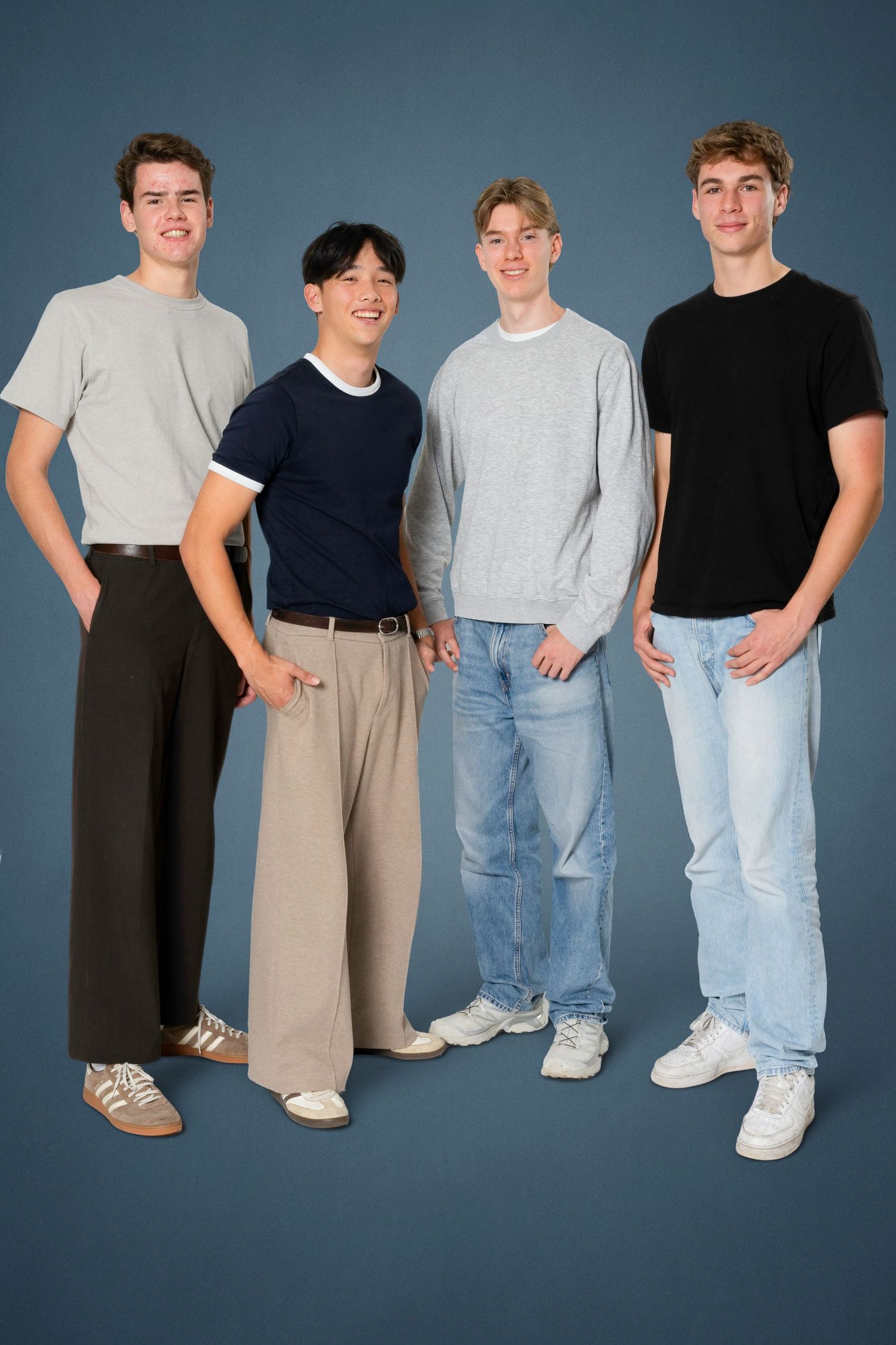 Four young men in casual outfits stand smiling together against a blue studio backdrop, wearing tees, a sweater, jeans and trousers.