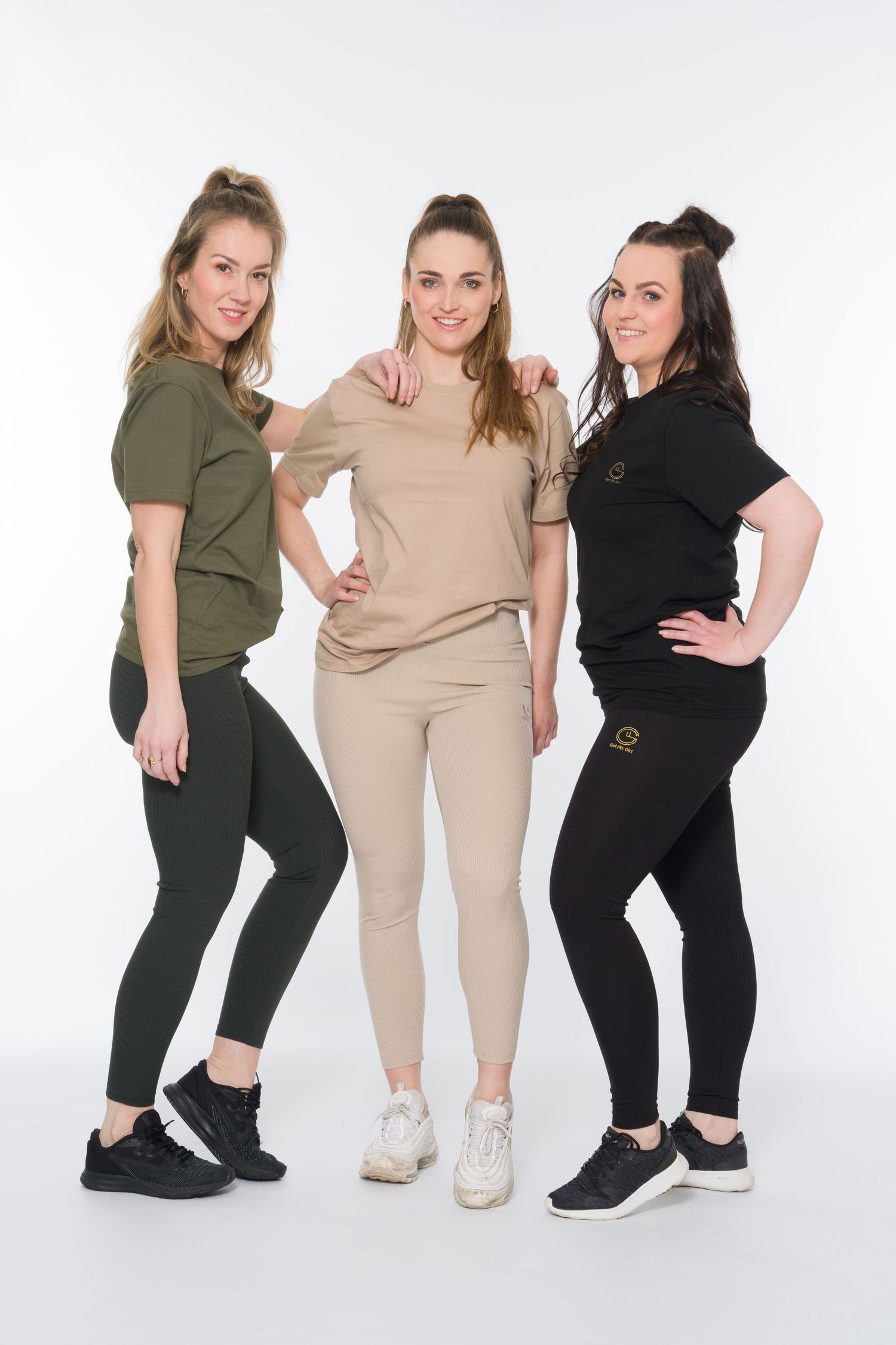 Three women in matching athletic wear posing together against white background in fitness studio.