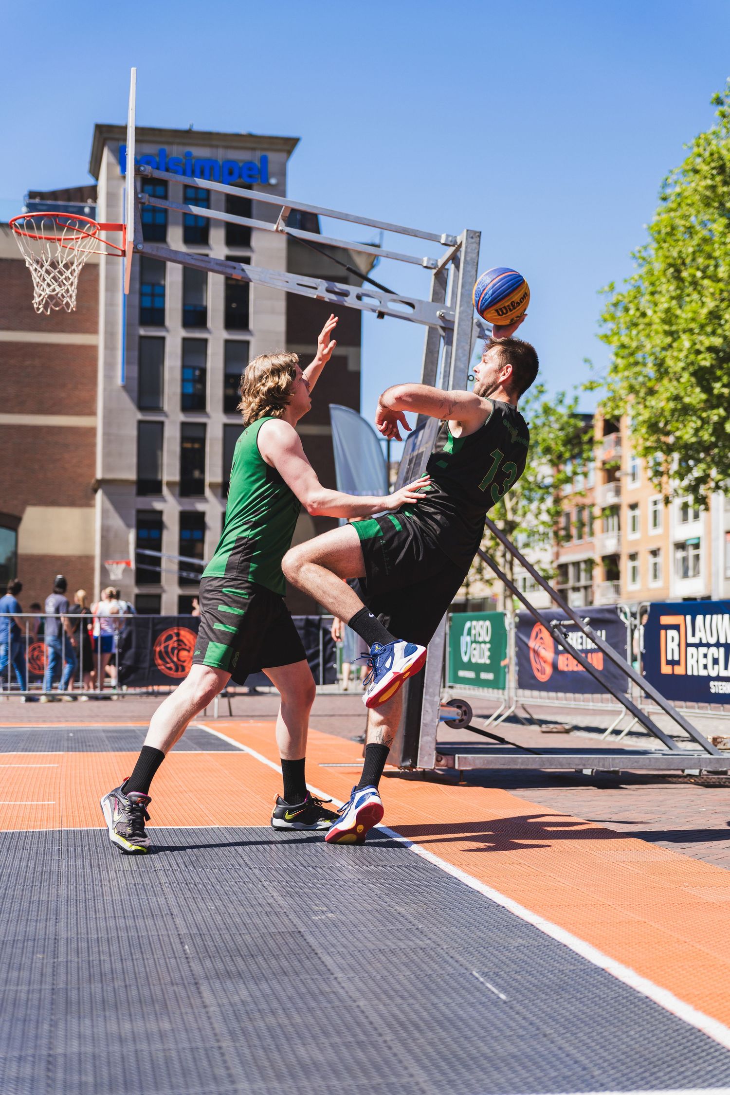 Two men playing street basketball outdoors, one jumping to block a shot near the hoop on a sunny day.