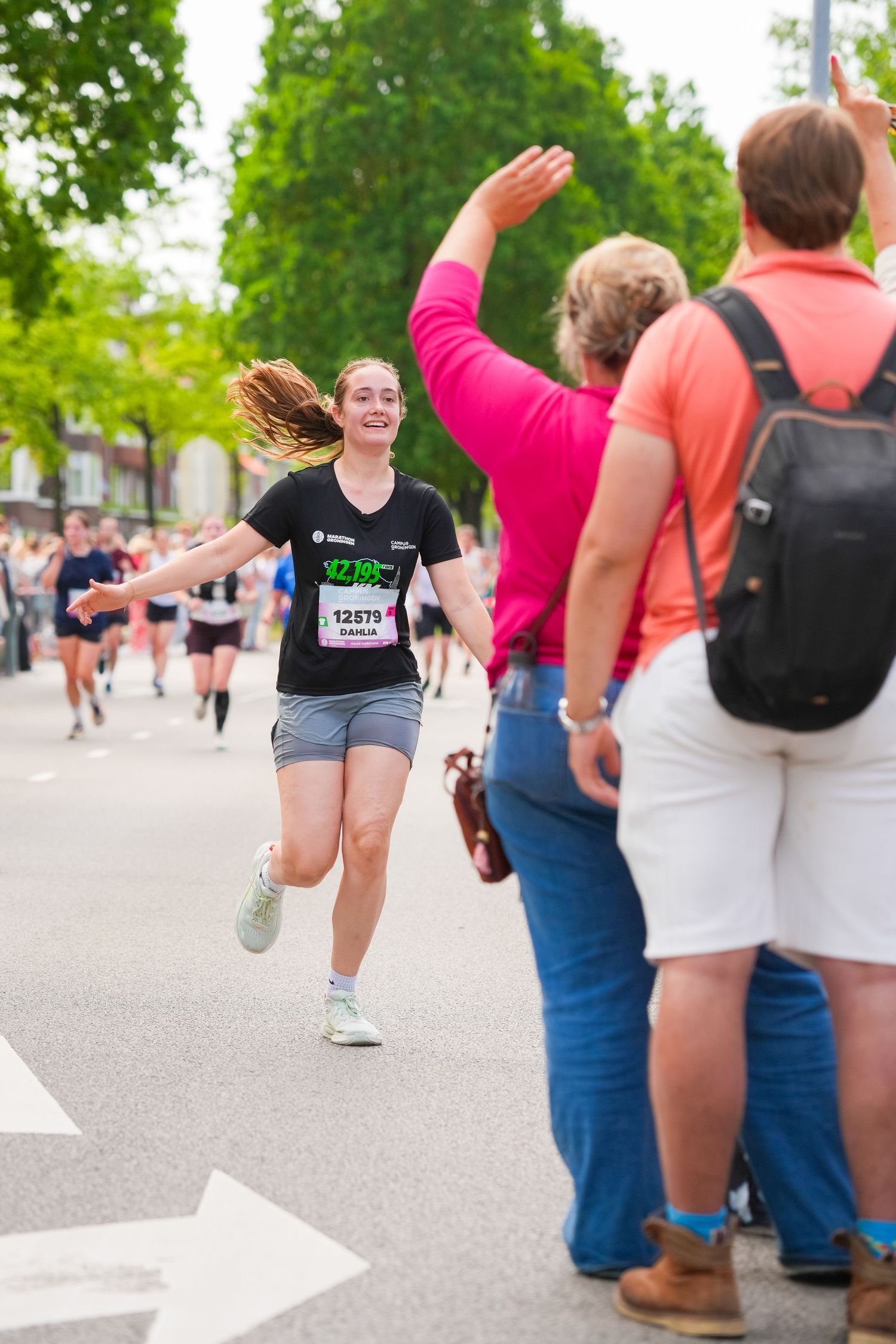 Smiling female marathon runner high-fiving spectators on a tree-lined race course during a road running event.